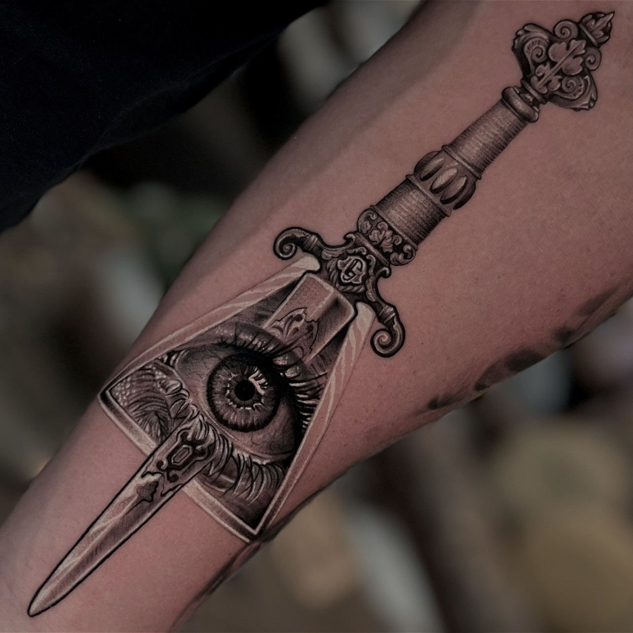 Tattoo of a sword with a detailed handle, featuring an eye with a realistic iris inside the blade of the sword.