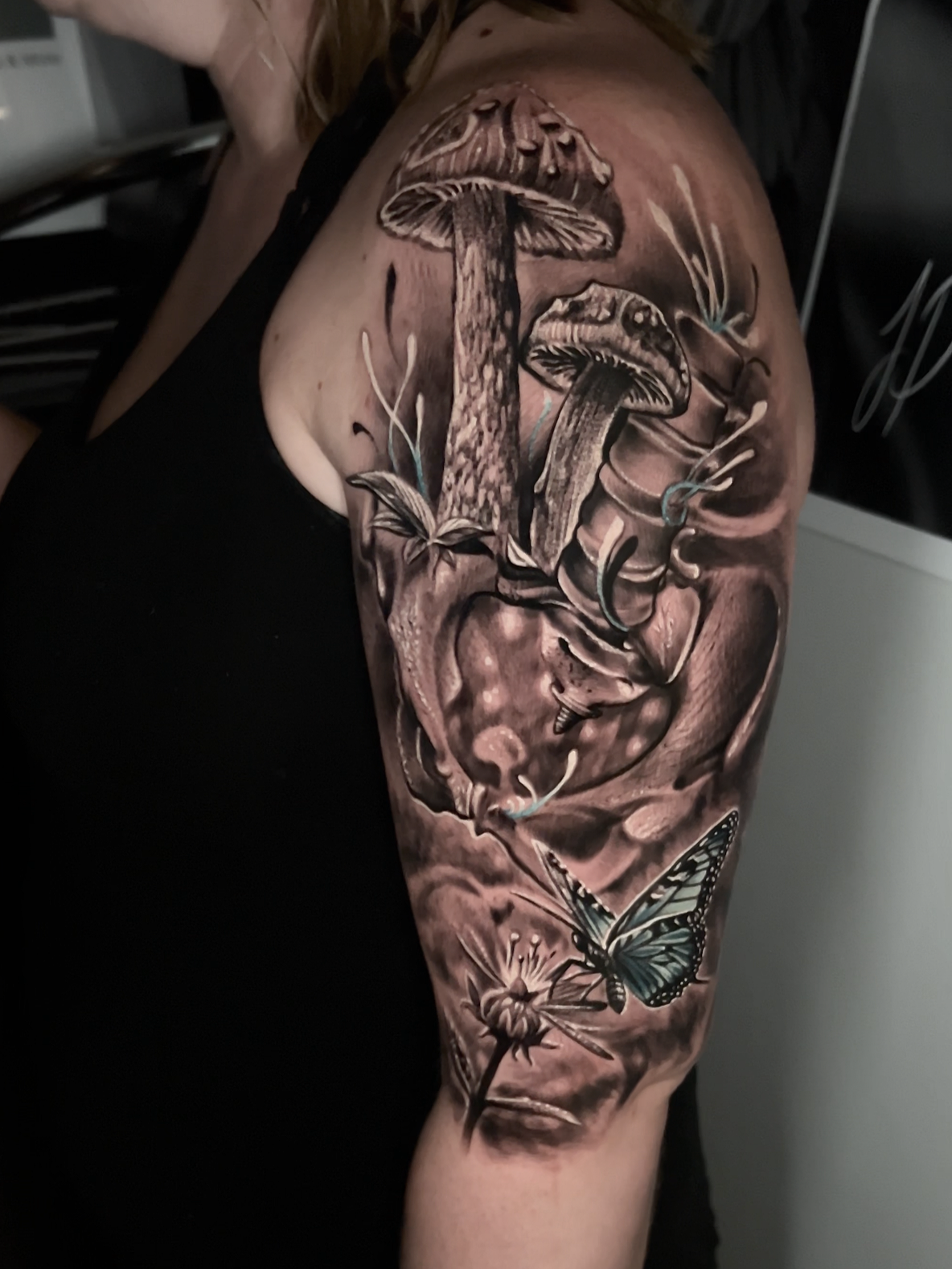 Black and gray tattoo of three mushrooms, a butterfly, and dandelions on a person's upper arm and shoulder.