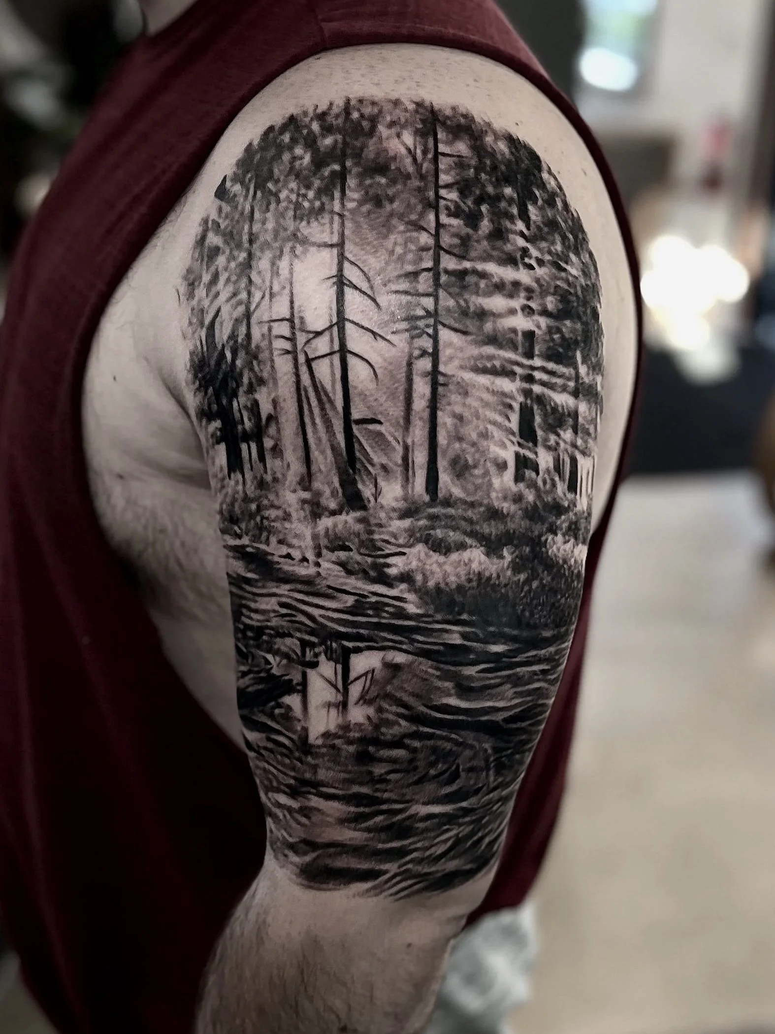 Black and gray forest landscape tattoo on person's upper arm, featuring trees and a body of water.