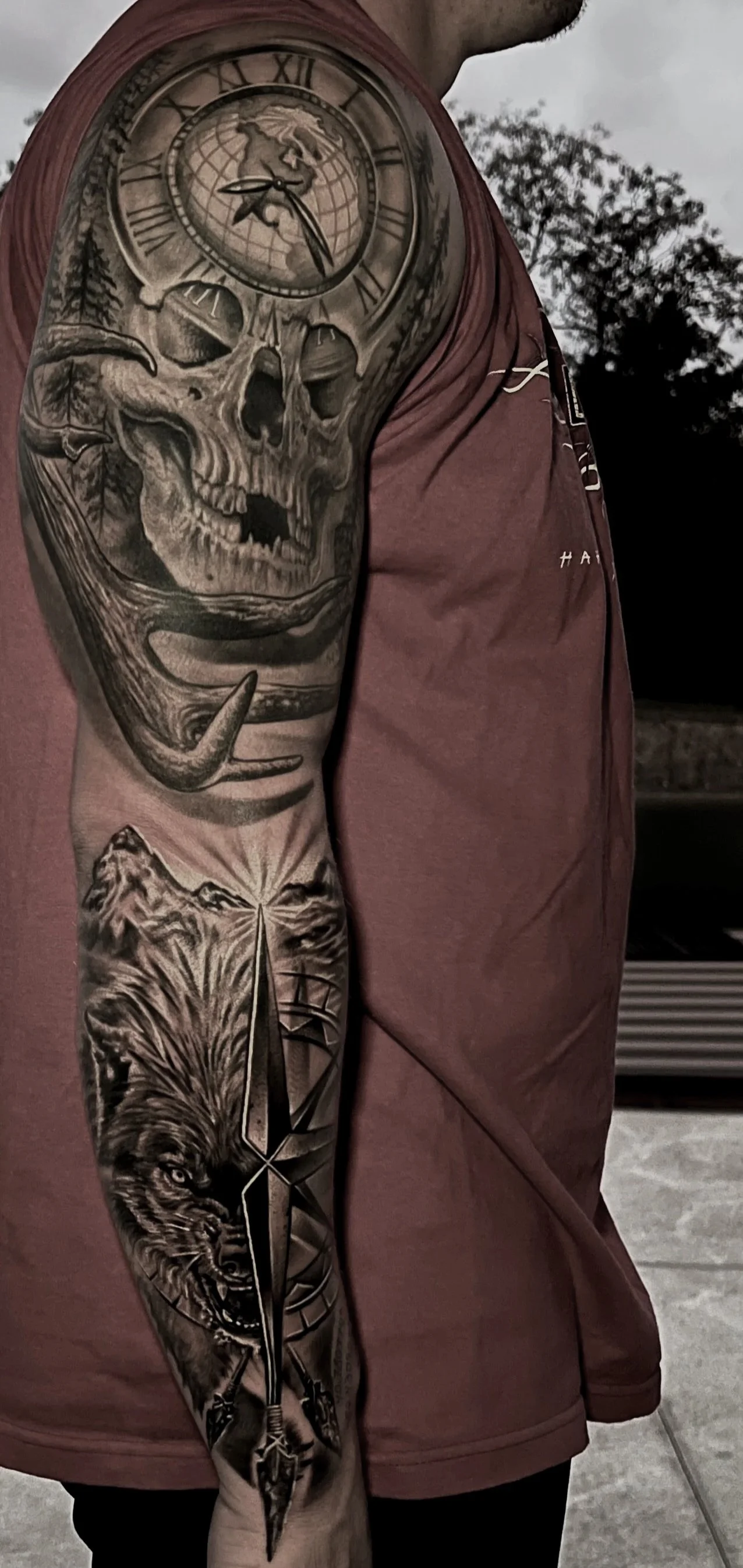 Tattoo sleeve on man’s arm featuring a skull with a clock on top, a howling wolf with a dagger, and mountains in the background.