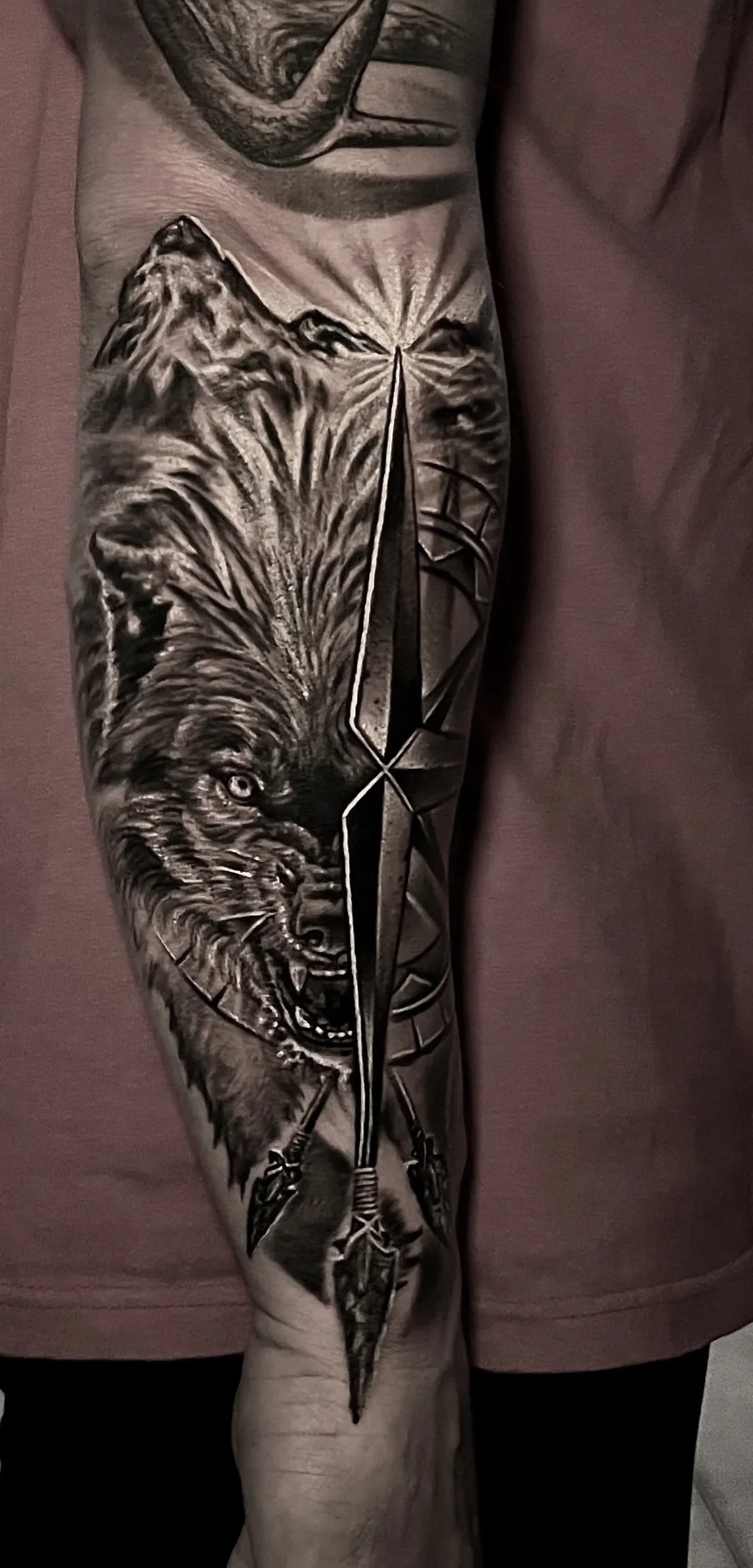 A detailed black and gray tattoo of a wolf's head howling, with a lone arrow piercing through the middle, on a person's forearm.
