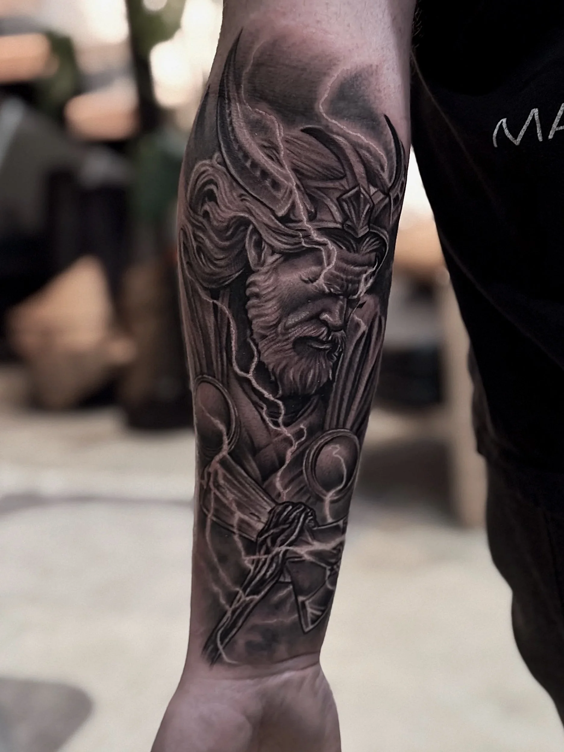 Black and gray tattoo of a mythological warrior with a beard, helmet, and long flowing hair, holding a staff or spear.