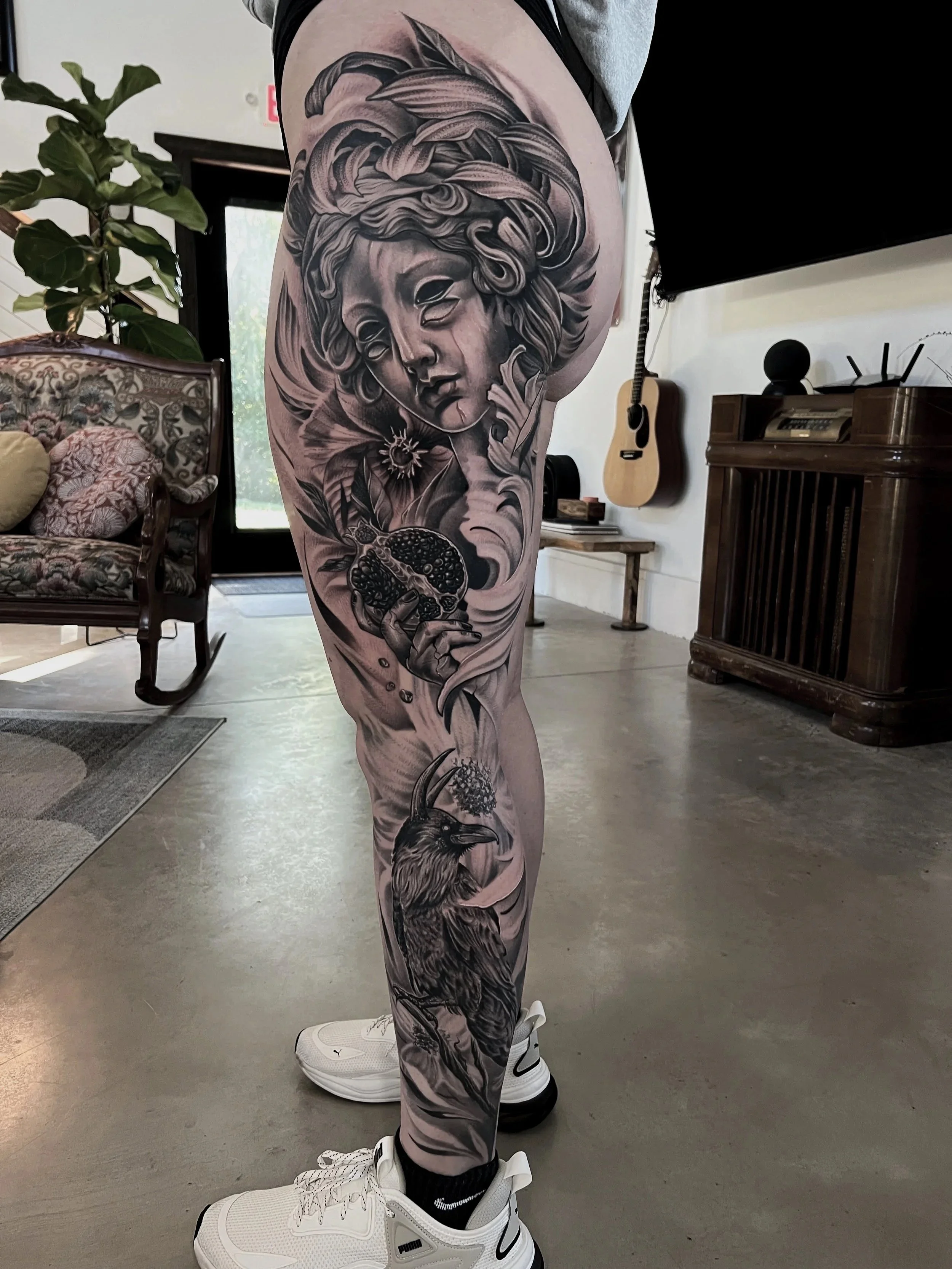 Tattoo of a female face with flowing hair, a flower, a hand holding a pomegranate, and a bird on the thigh.