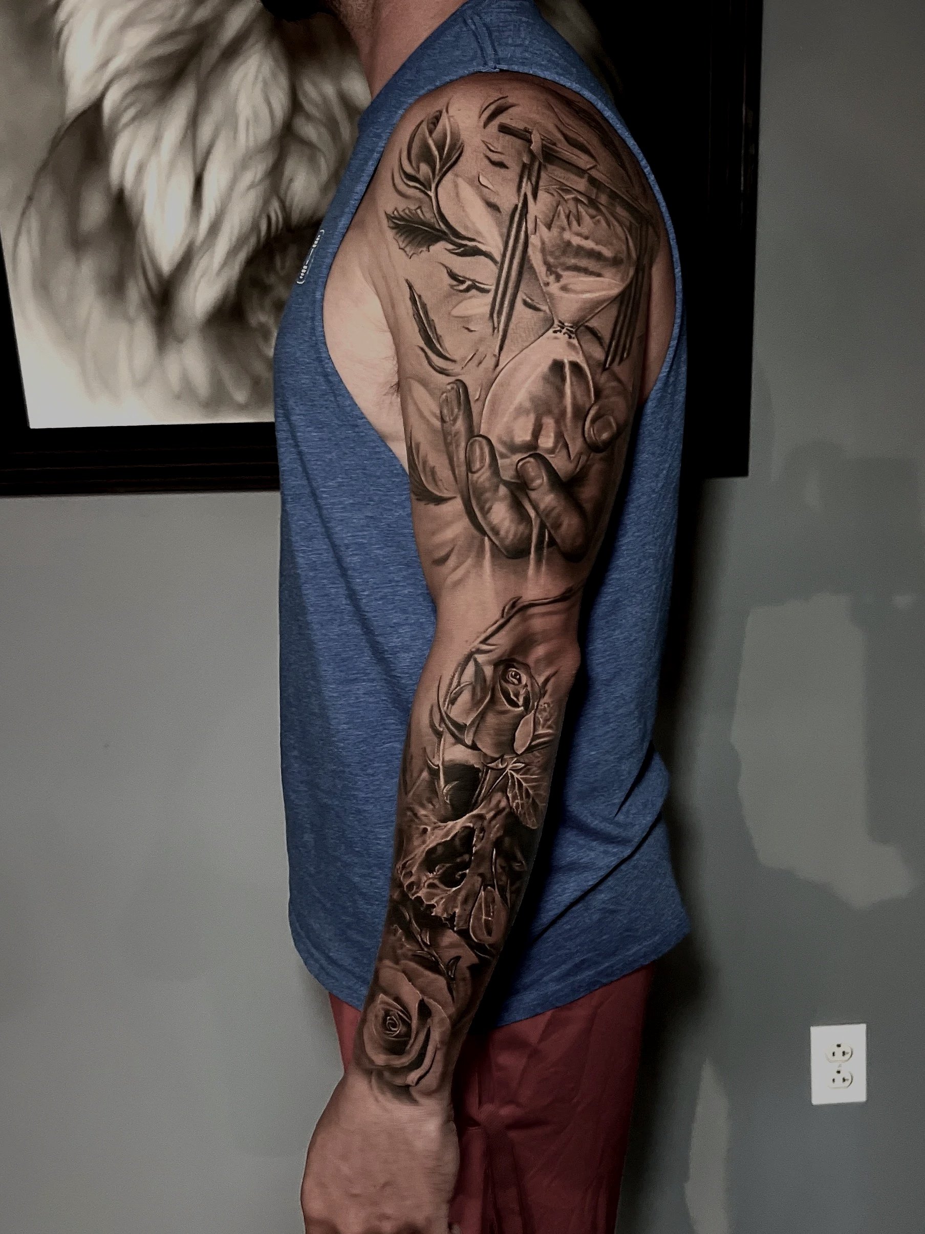 Black and gray realistic tattoo sleeve featuring a clock, a woman's face, a rose, and animal skulls, covering the upper arm and forearm of a man in a blue sleeveless shirt.