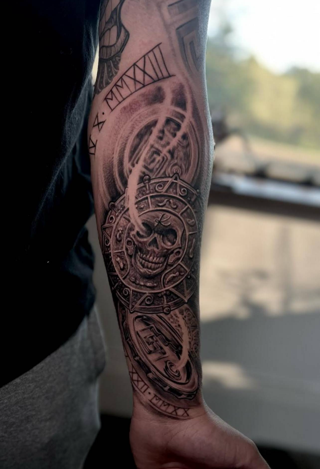 Close-up of a person's upper arm with a detailed black and gray tattoo featuring a skull, clock gears, Roman numerals, and intricate designs.