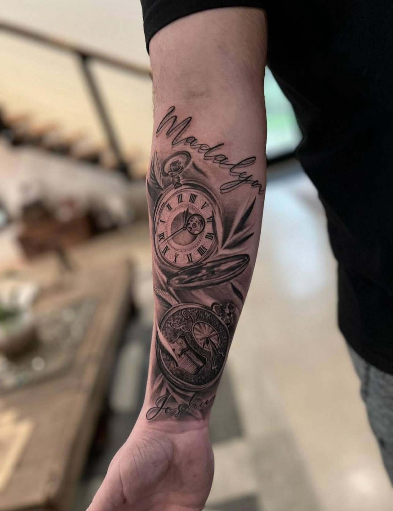 Black and gray tattoo of a pocket watch with Roman numerals, surrounded by leaves, with the words 'Madelyn' and 'Joshua' written in script.