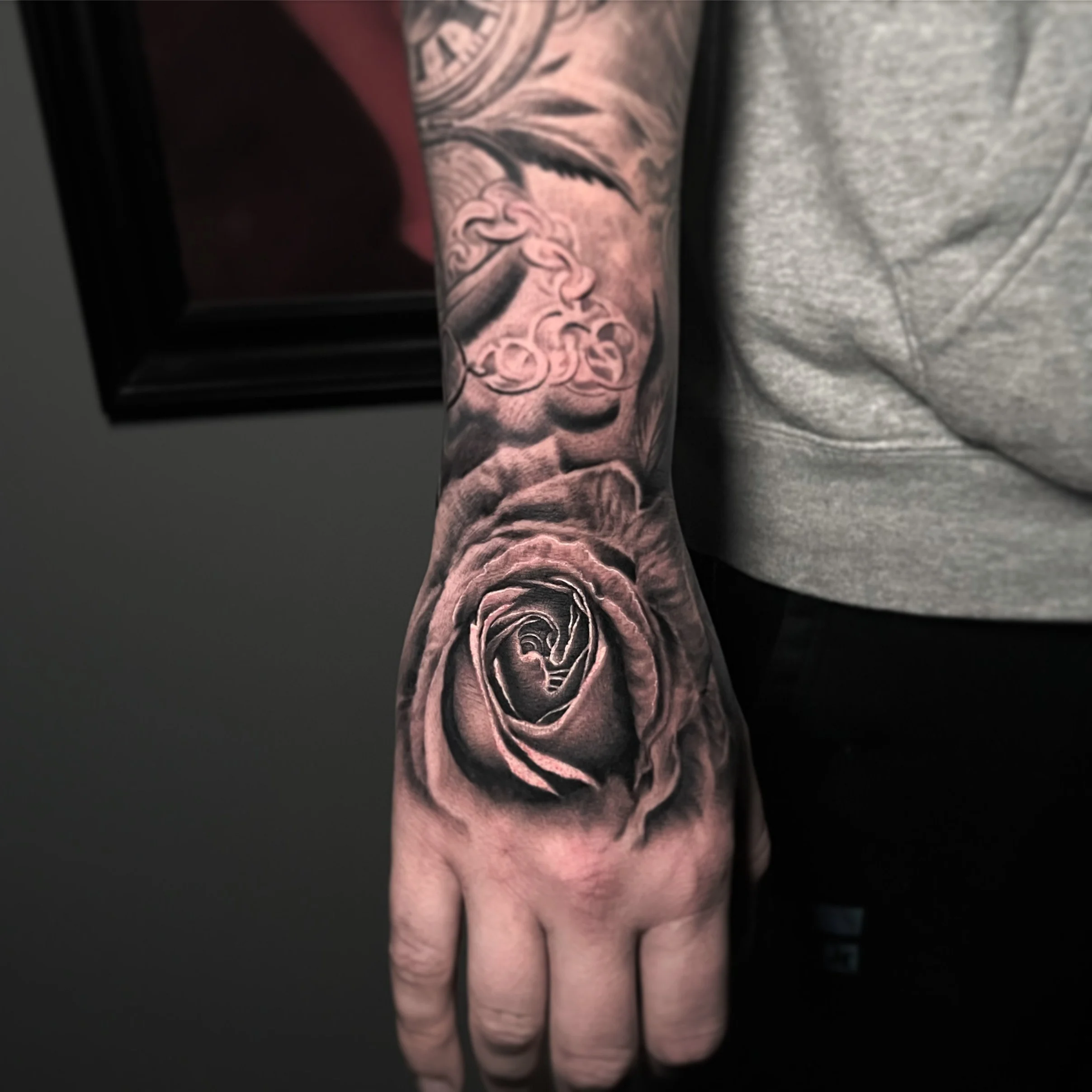 Close-up of a person's arm with a detailed black and gray tattoo of a rose on the hand and a floral design on the forearm.