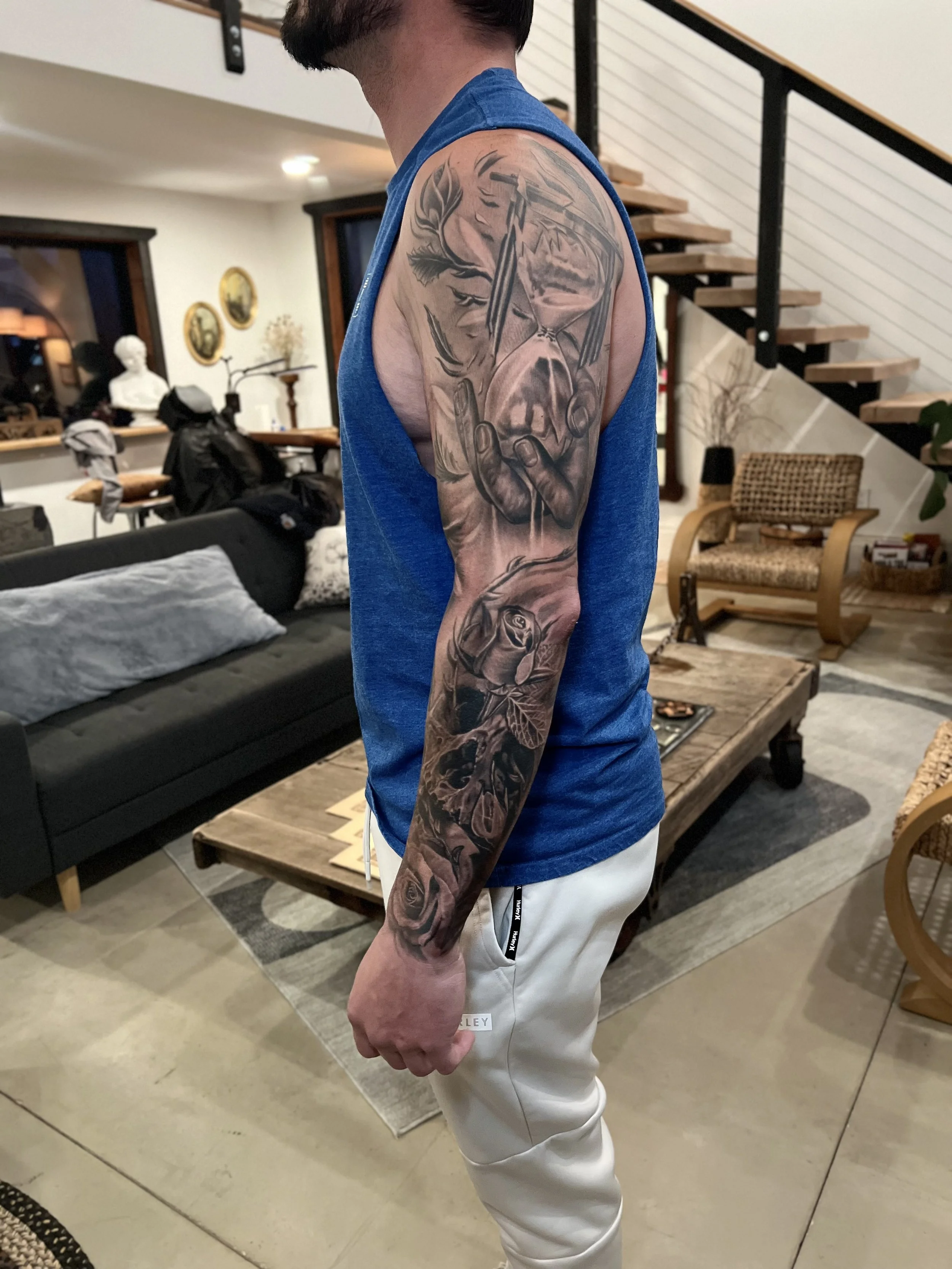 A man with a detailed black and gray tattoo sleeve on his left arm. The tattoo features a hand holding a knife with a face and a rose below.