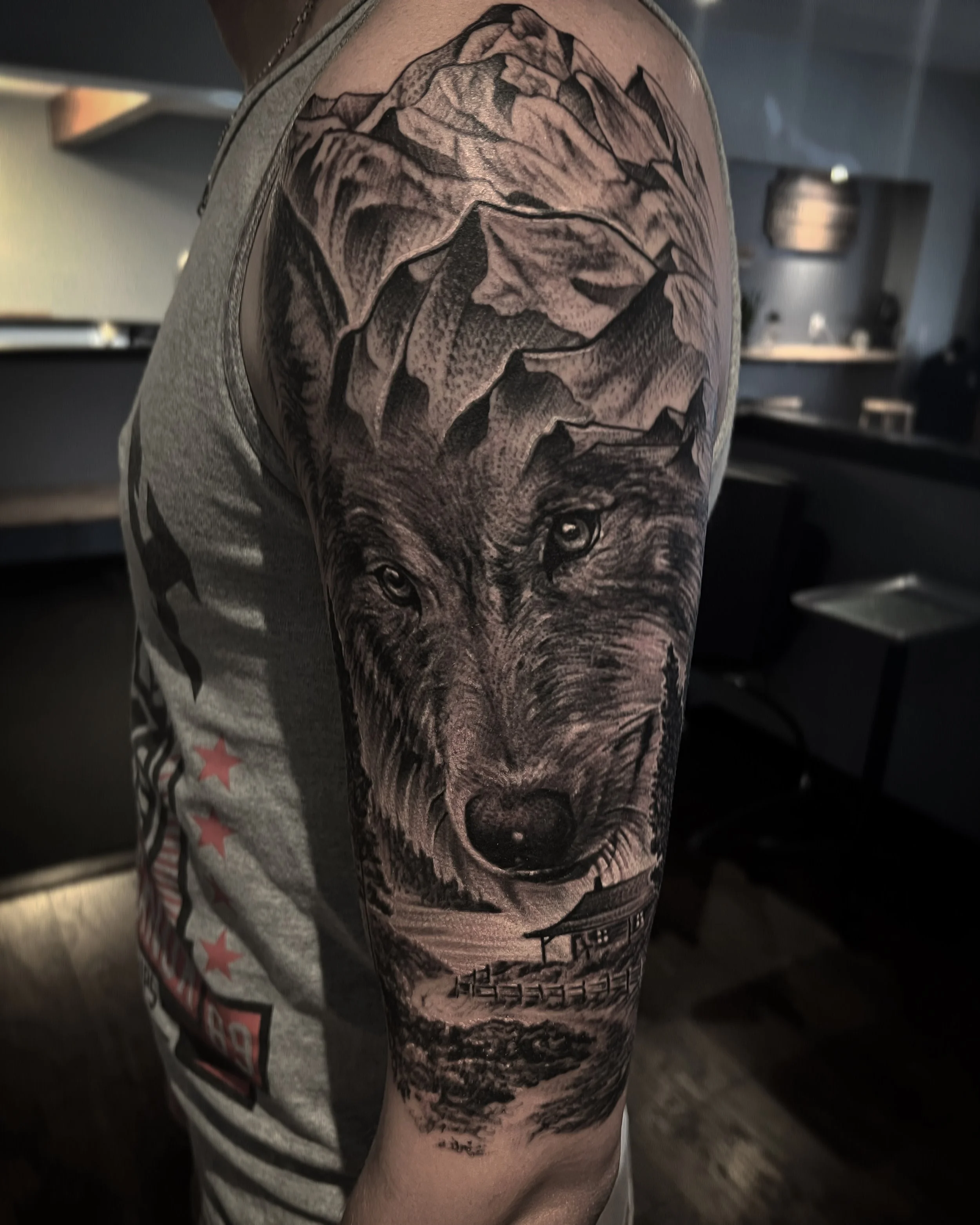 A detailed black and gray tattoo of a wolf's face combined with a mountain landscape and a wooden cabin on a person's upper arm.