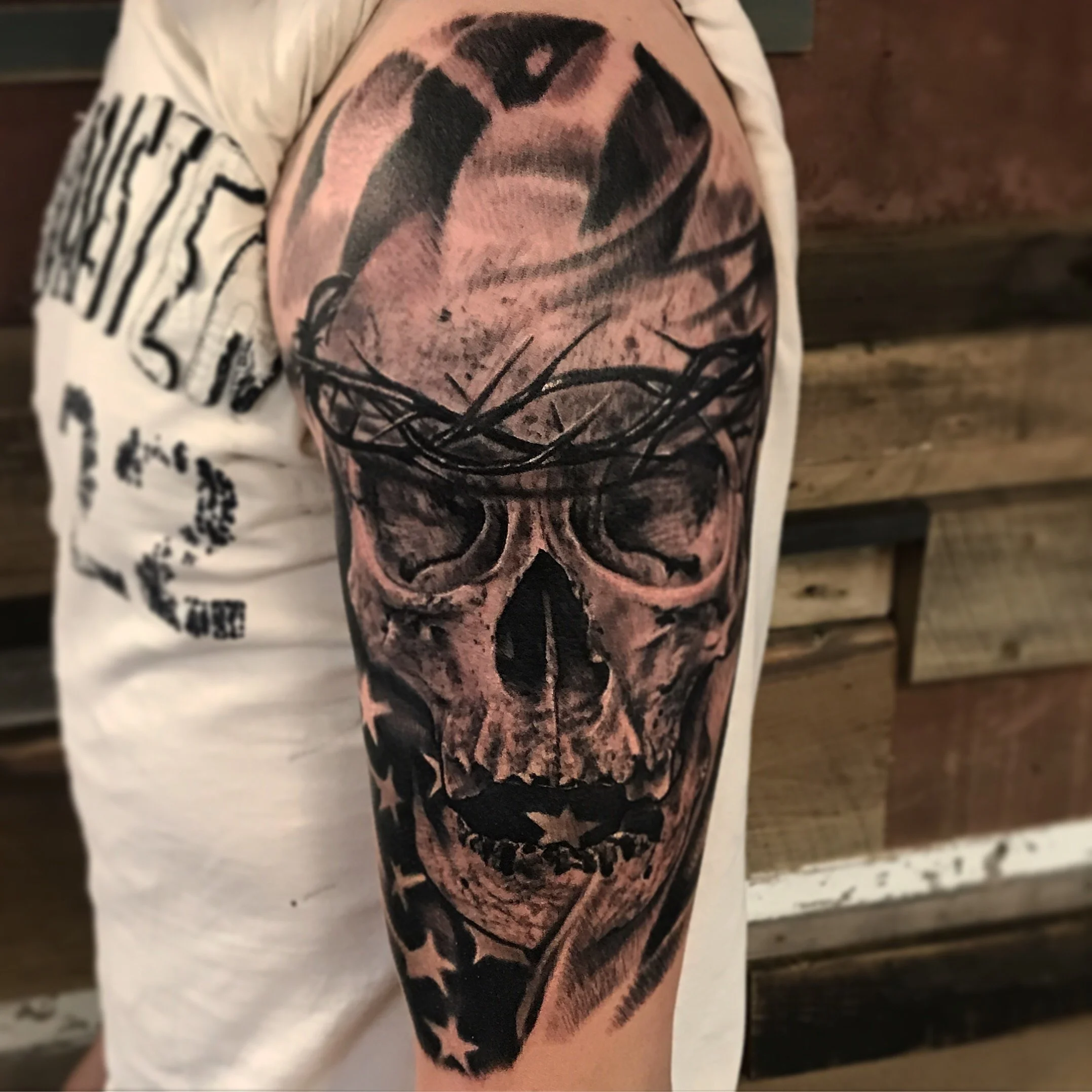 Tattoo of a realistic human skull with a crown of thorns, partially covered by an American flag, on a person's upper arm.