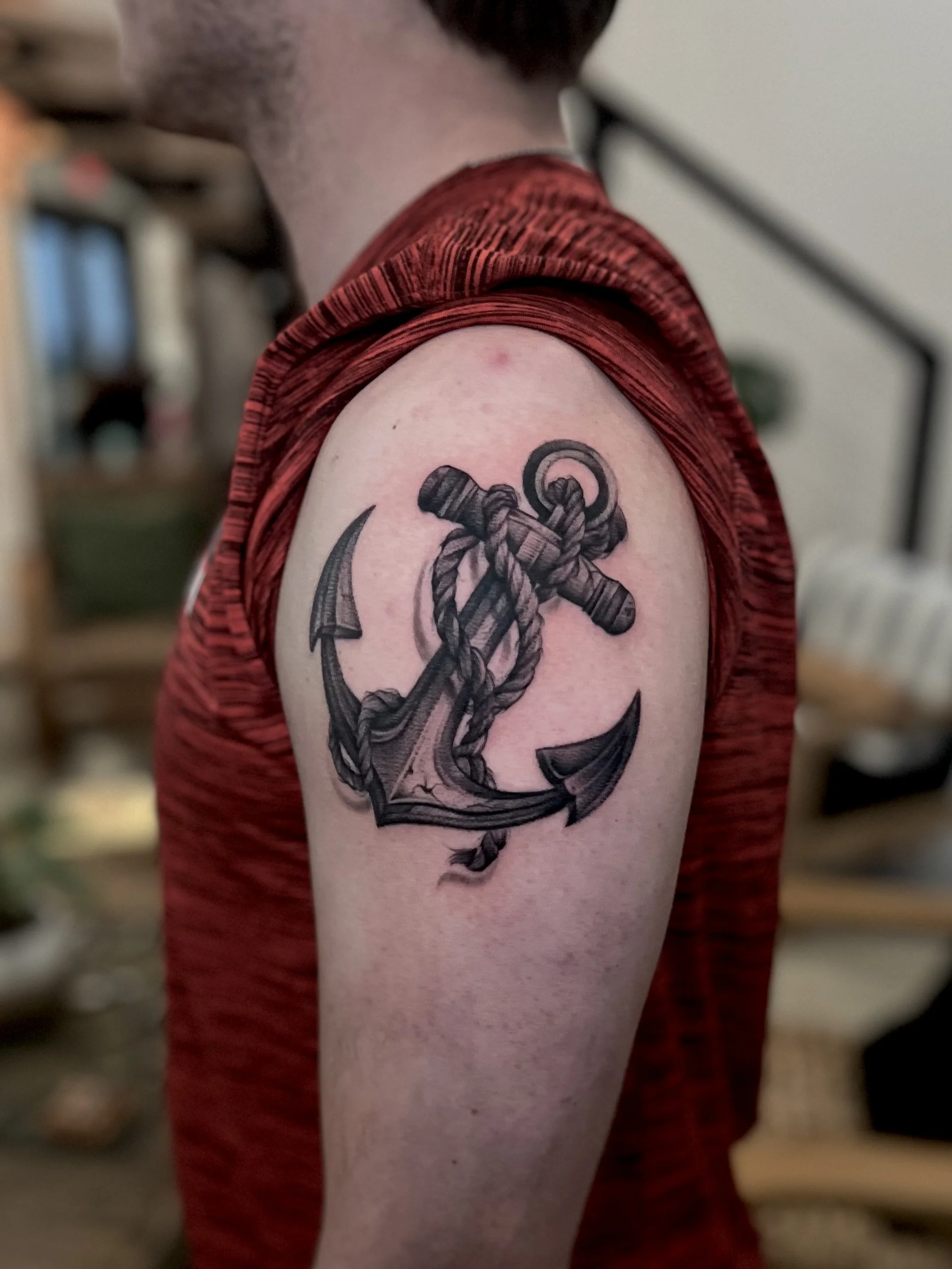 Black and gray tattoo of an anchor wrapped with a rope on the upper arm of a person wearing a red sleeveless shirt.