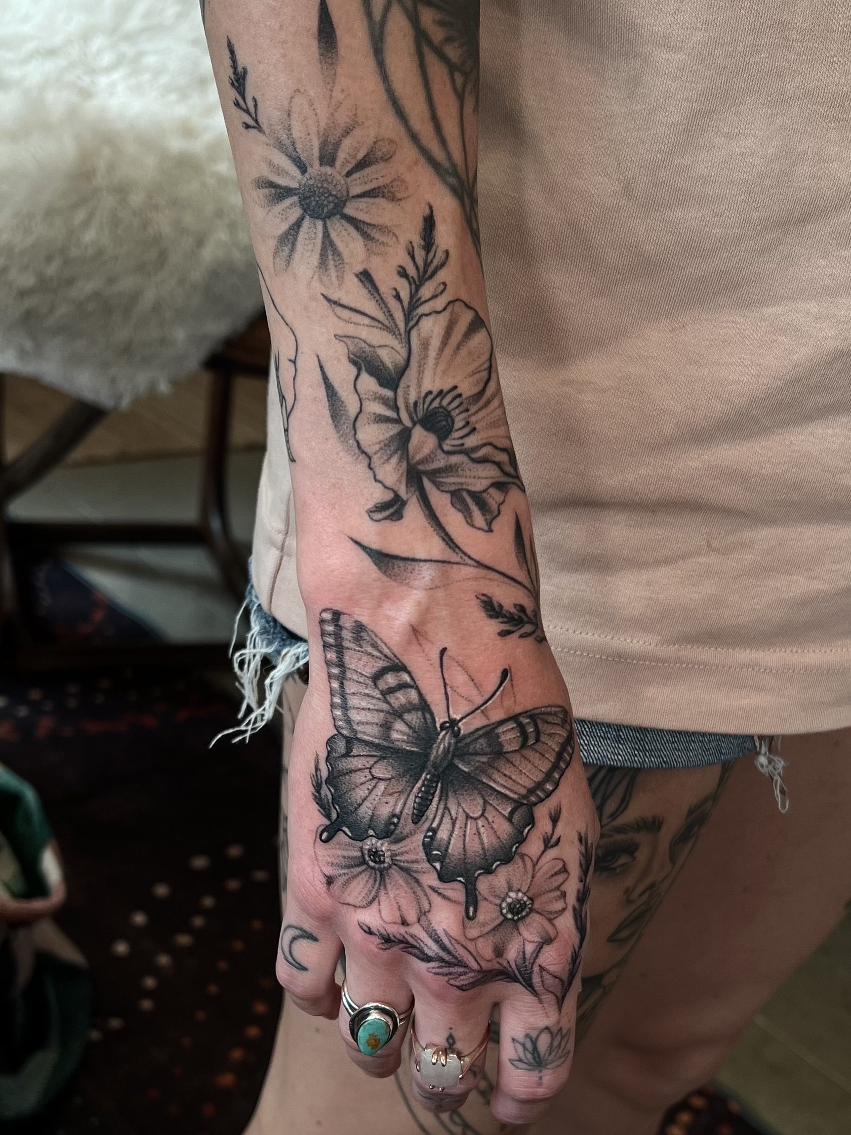 A person's hand and wrist with multiple black and grey floral and butterfly tattoos, wearing rings, including one with a turquoise stone, and a crescent moon tattoo on the finger.