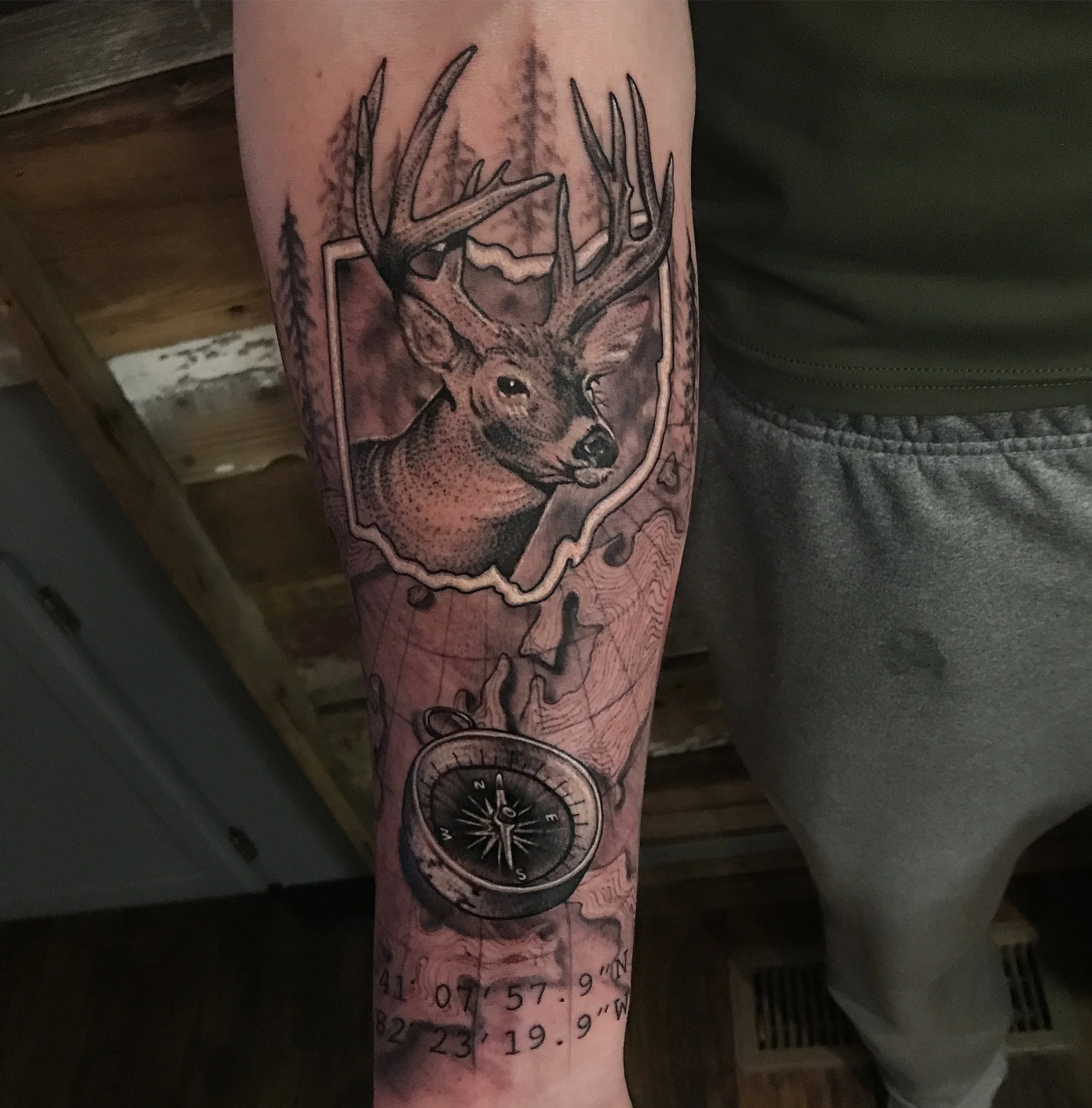 Tattoo of a deer head with antlers, map background, and a compass on the arm.