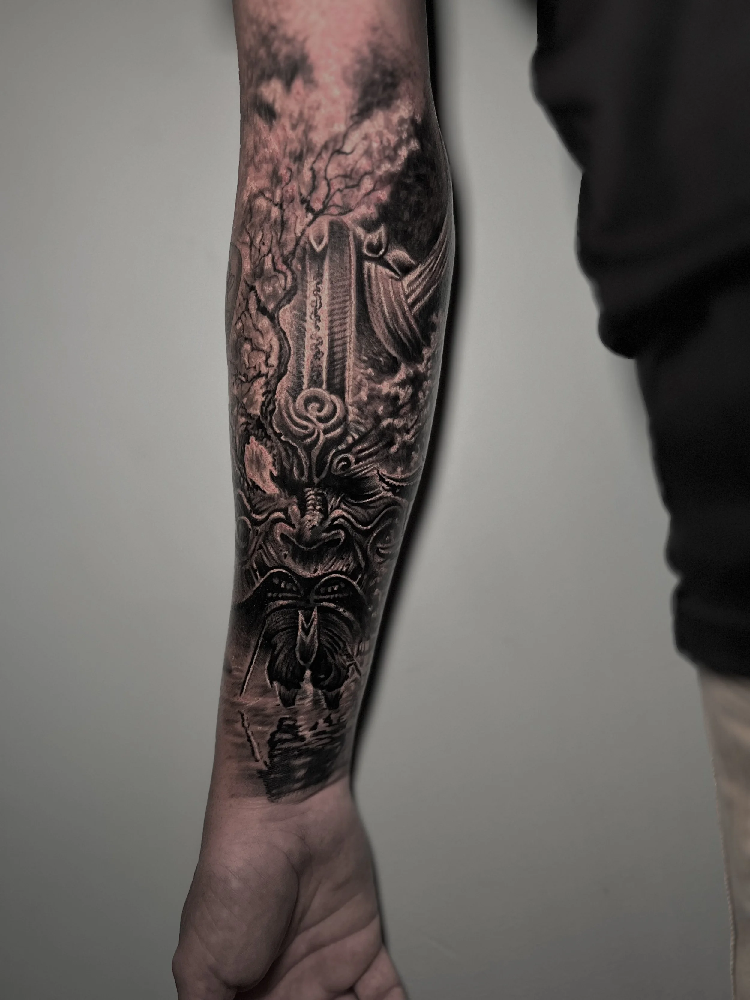 Black and gray tattoo of a mythological creature with an angry face and horns, standing in front of a stormy sky with clouds, on a person's forearm.