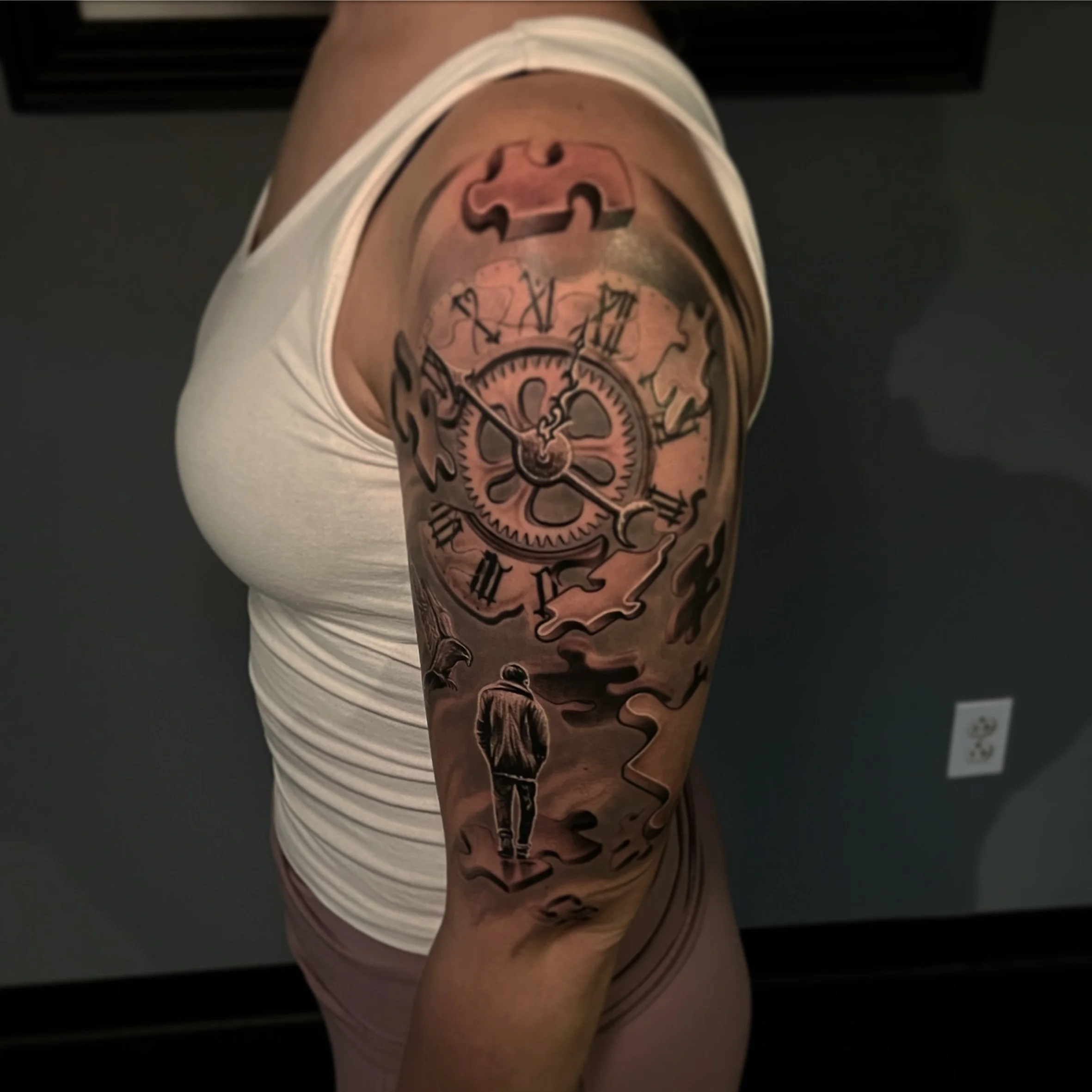 Tattoo of a clock with Roman numerals and mechanical gears, a puzzle piece at the top, and a man walking on a path beneath the clock on a person's upper arm.