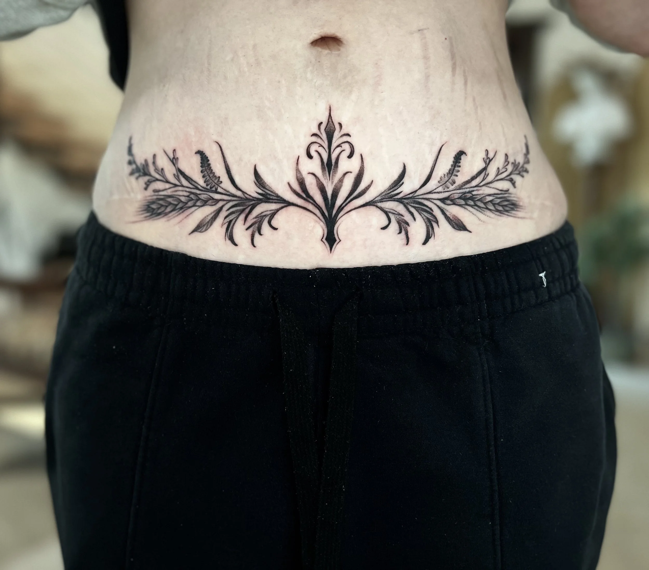 A tattoo of a symmetrical, ornamental design resembling a stylized plant or floral pattern, centered on the lower abdomen near the waist, with black ink on light skin, paired with black pants.