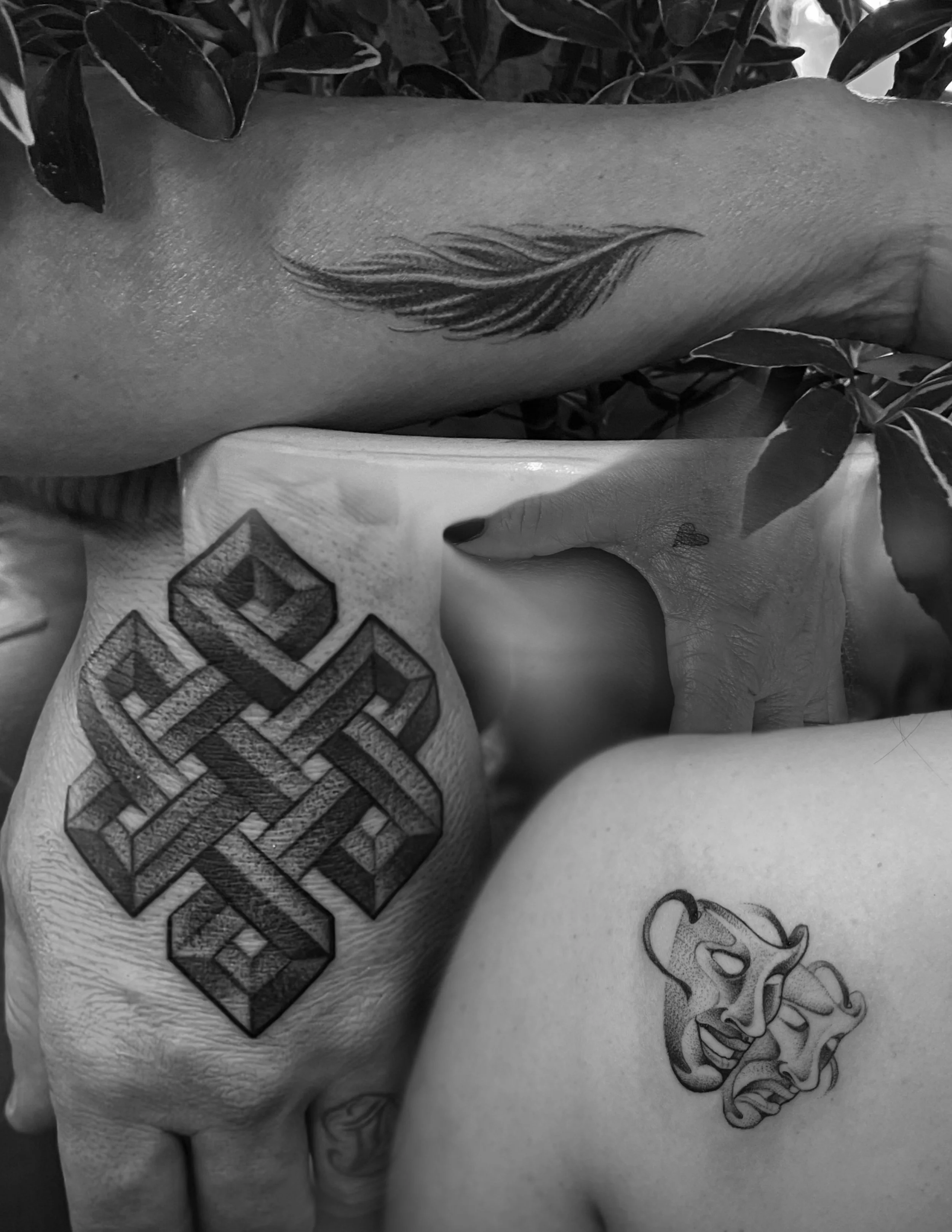 Black and white photo of three tattoos on intertwined human limbs, with one arm showing a feather tattoo, a large geometric cross tattoo on another arm, and two theatrical mask tattoos on a third limb, surrounded by leaves.
