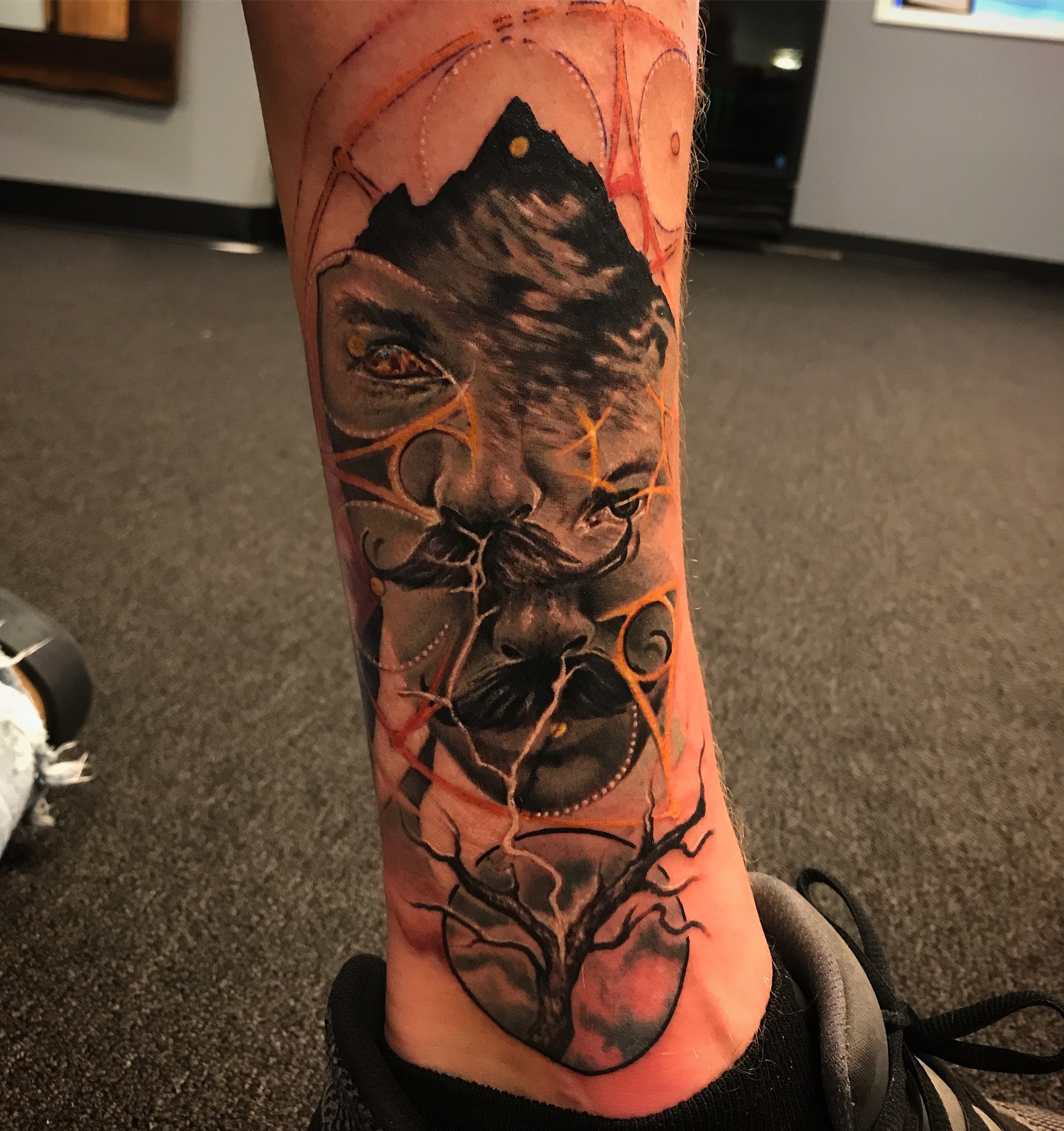 Realistic tattoo of a man's face with mustache and beard, with a mountain and sky in the background, intertwined with orange and black abstract lines and shapes.