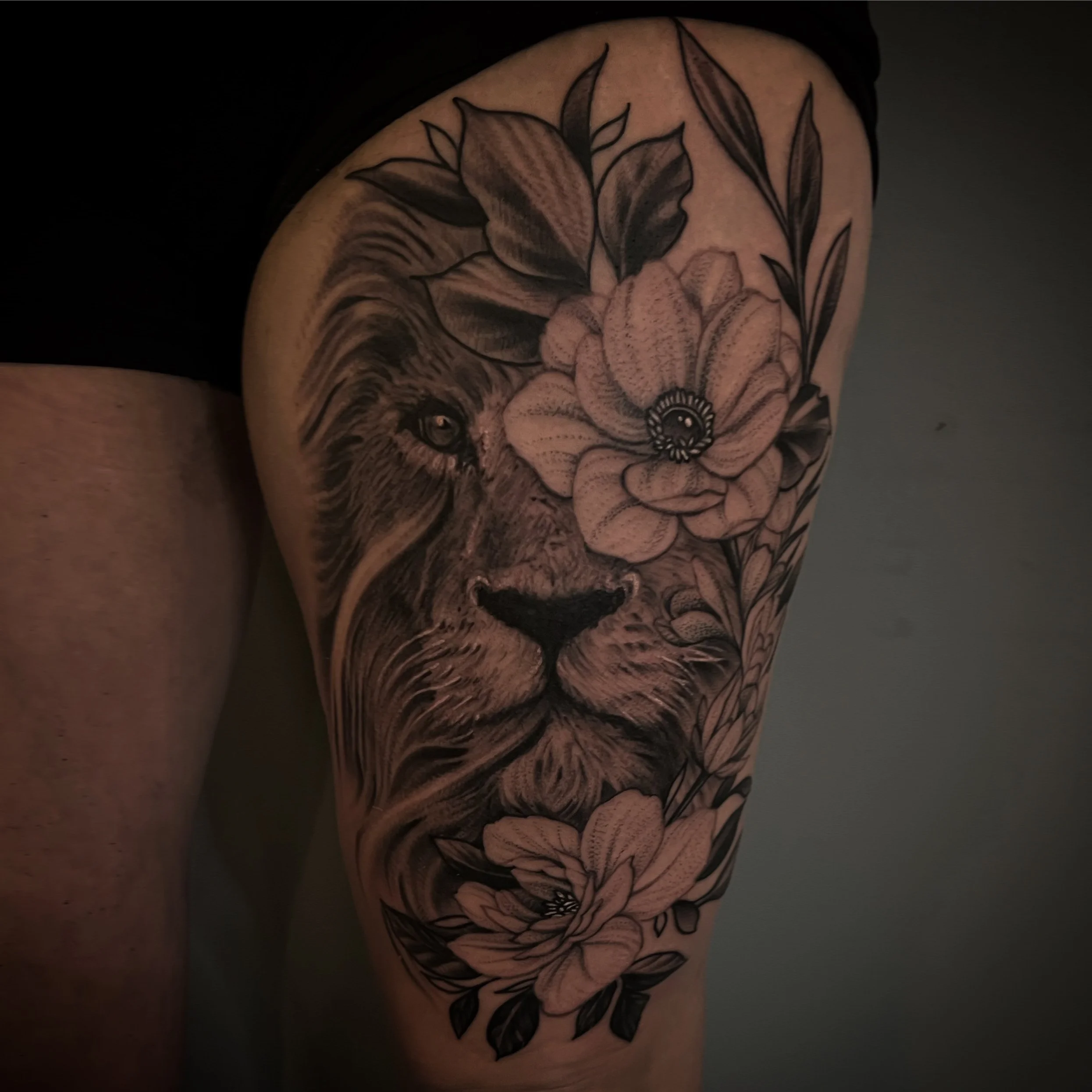 Black and gray tattoo of a lion's face surrounded by large flowers and leaves on a person's upper arm.