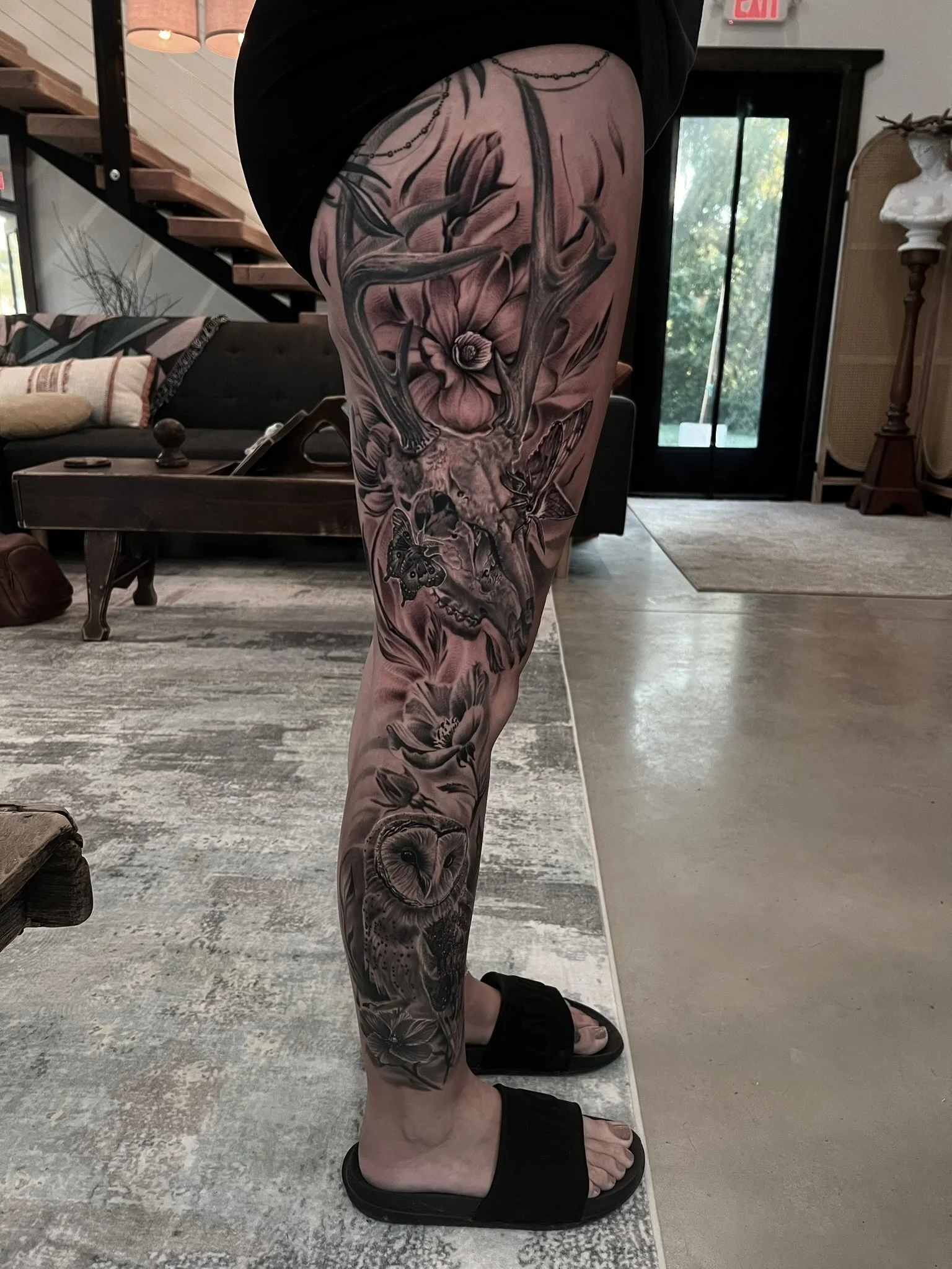 Tattooed leg with floral and owl designs in black and gray ink, wearing black slide sandals.