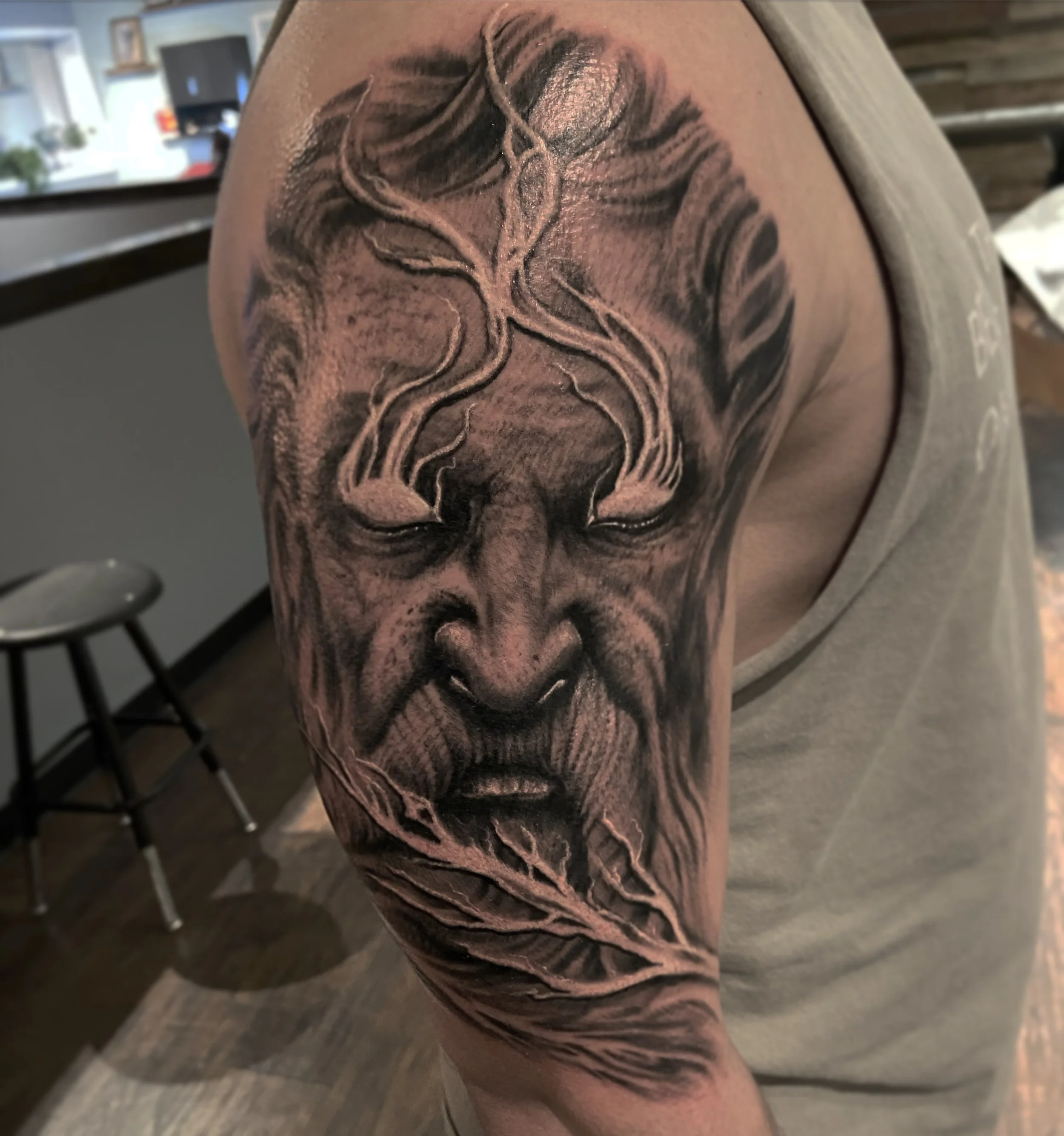 Black and gray tattoo of a bearded man's face with closed eyes and flowing hair, on a person's upper arm.