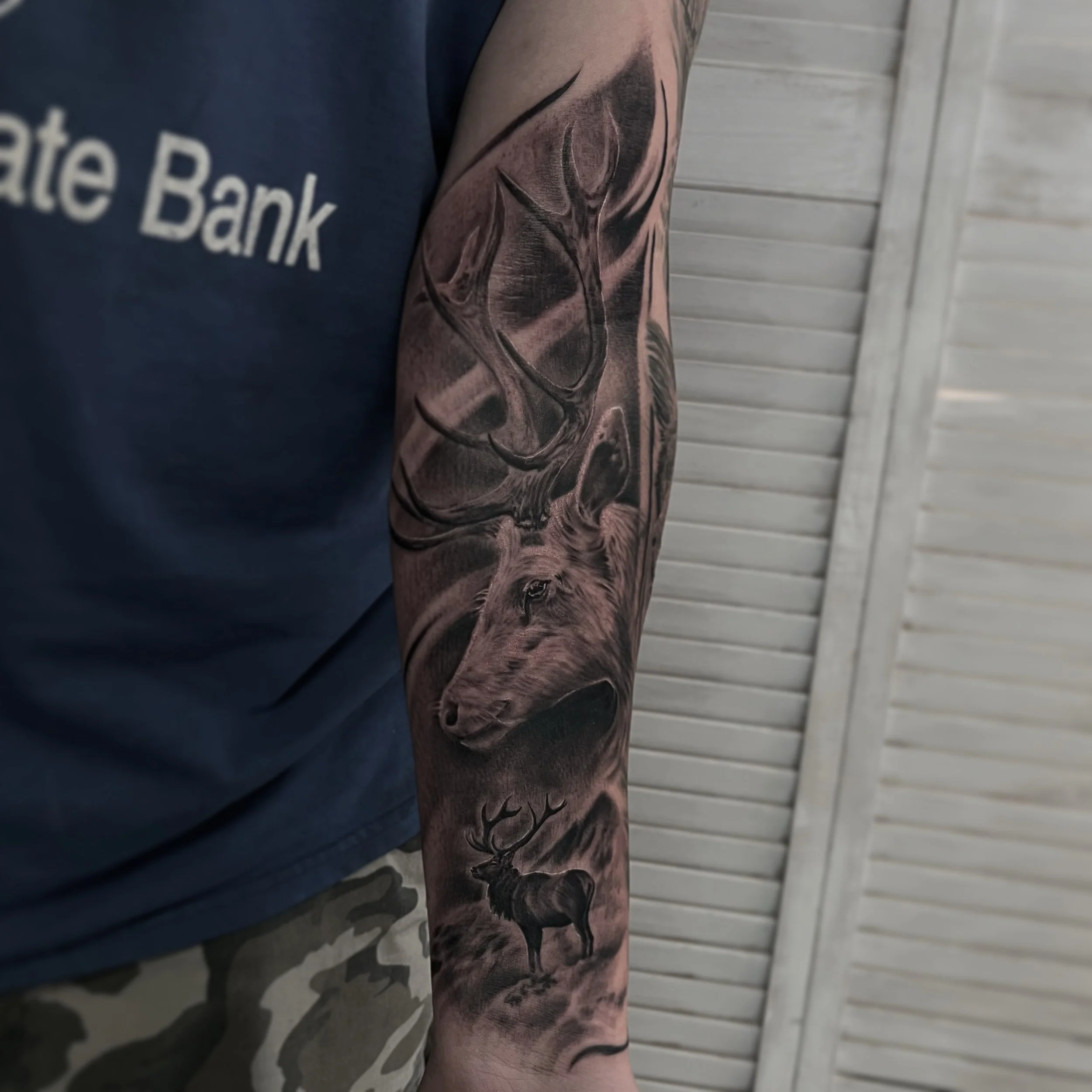 Black and gray realistic tattoo of a wolf and a stag on a person's forearm.