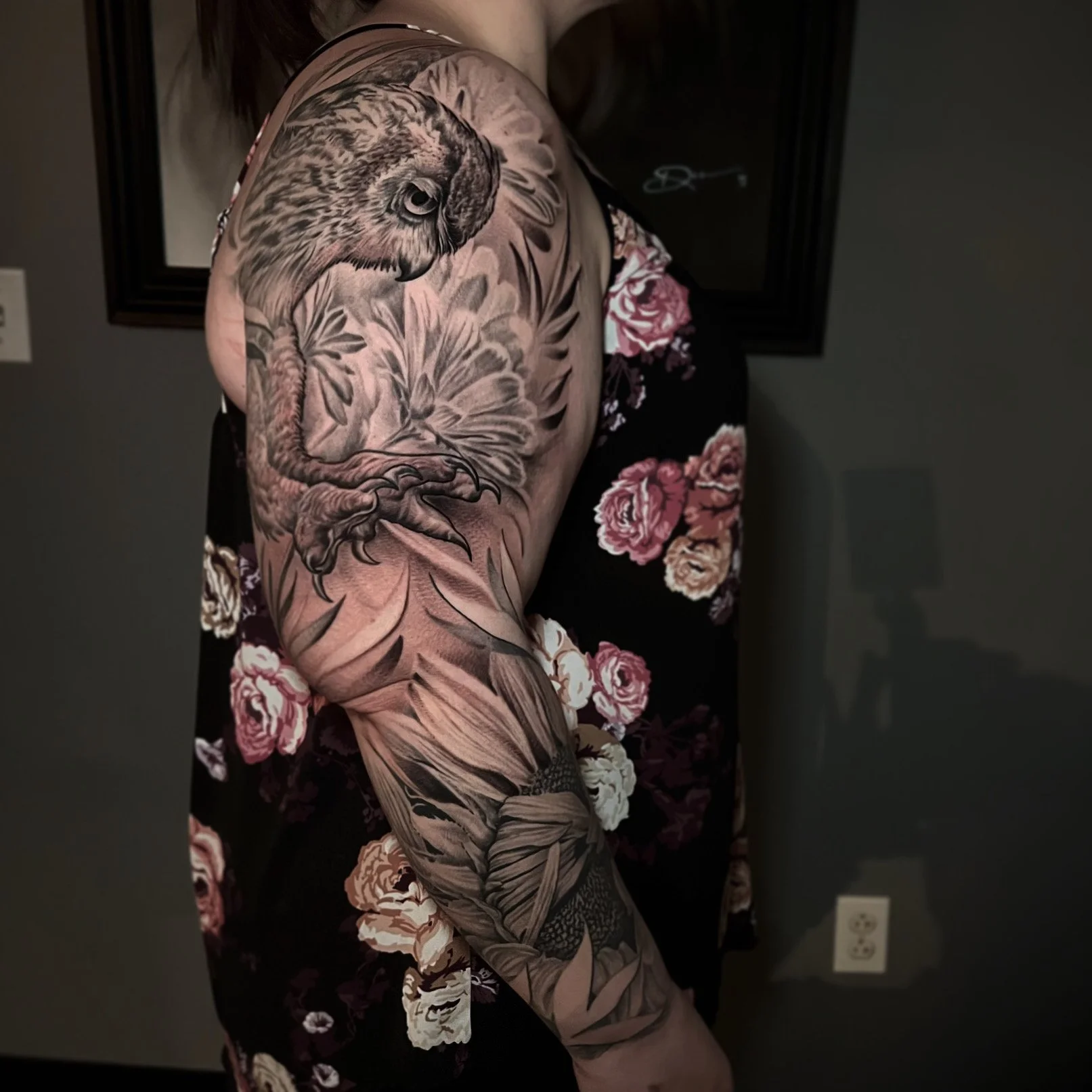 Close-up of a person's arm with a detailed black and gray tattoo of an owl perched on a branch, surrounded by leaves and flowers.