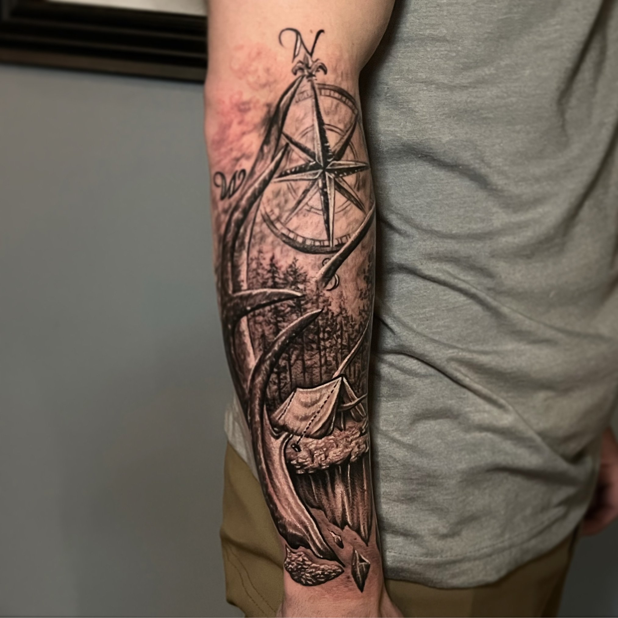 Black and gray tattoo of a compass, sailboat, and pine trees on a person's forearm.