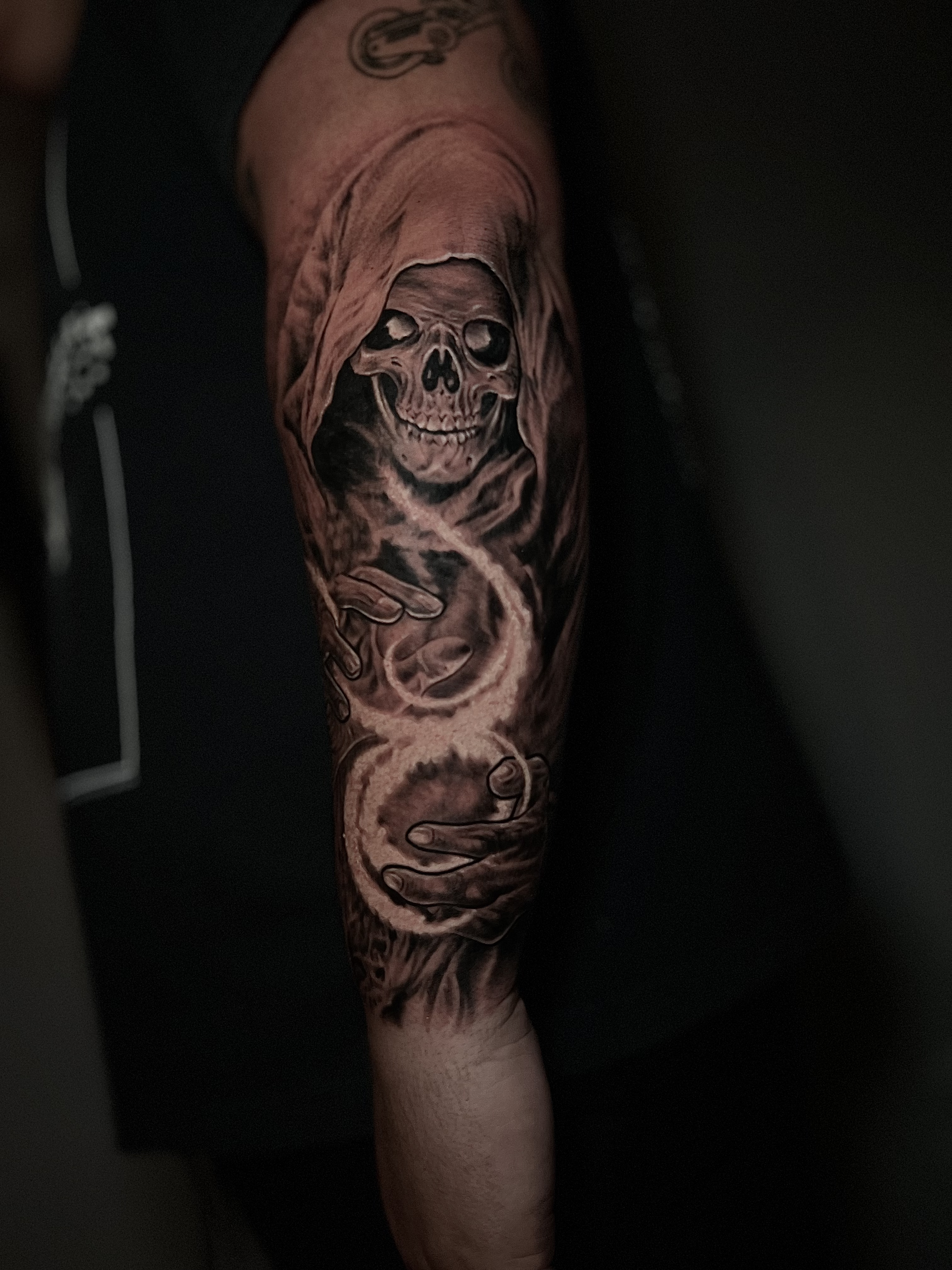 Tattoo of a skeleton with a hood and a smile, holding a scythe, on person's arm.