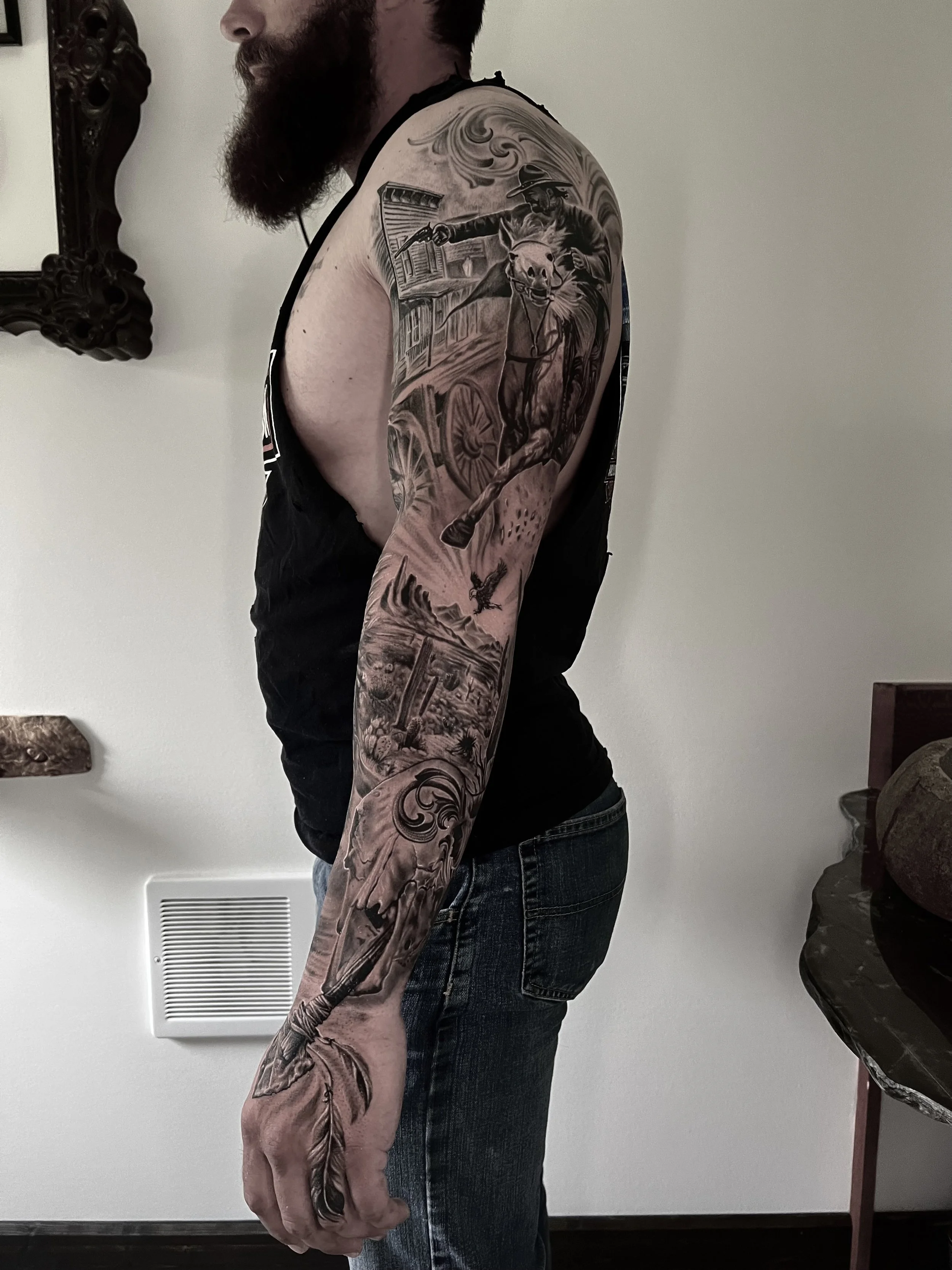 A man with a beard wearing a sleeveless black shirt and jeans, showing a detailed black and gray tattoo sleeve on his left arm, featuring a cowboy on a horse, a western scene with a building, an eagle, and feathers.