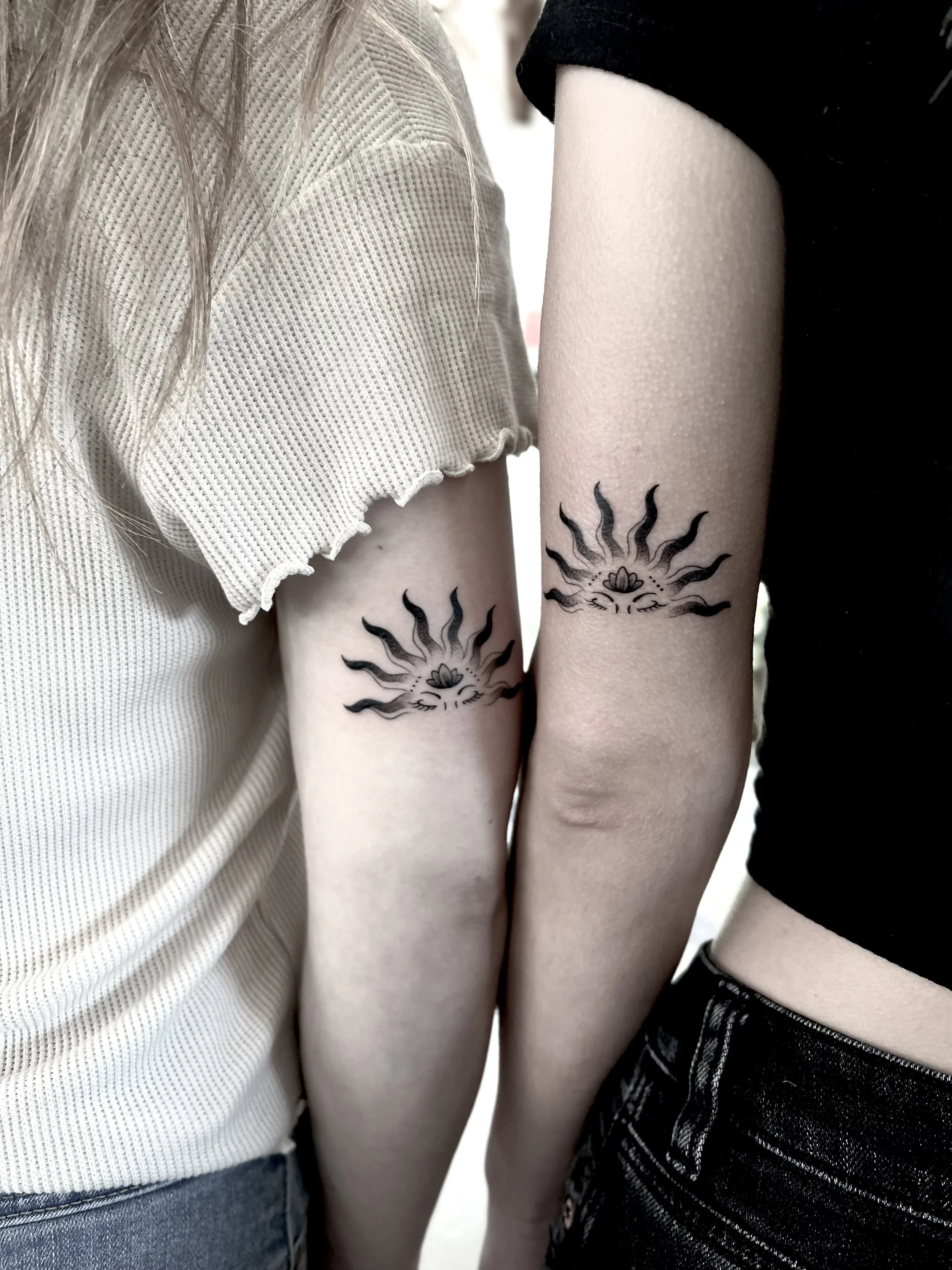 Close-up of two people standing side by side, showing their upper arms with matching black sun tattoos with wavy rays and a small central motif.