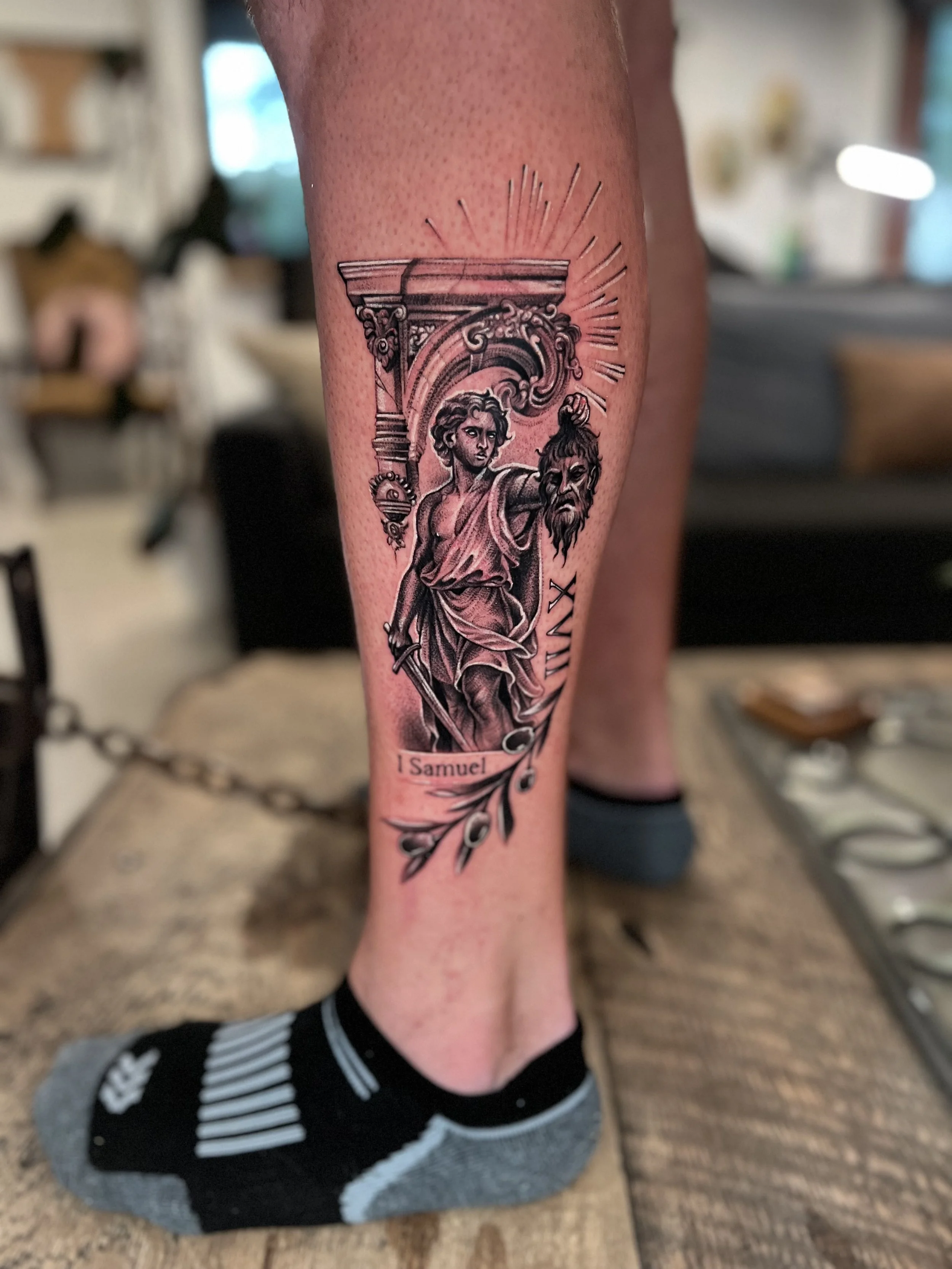 Black and gray tattoo on person's calf depicting a biblical scene with a woman holding a severed head, church architecture, and a banner with the text '1 Samuel'.