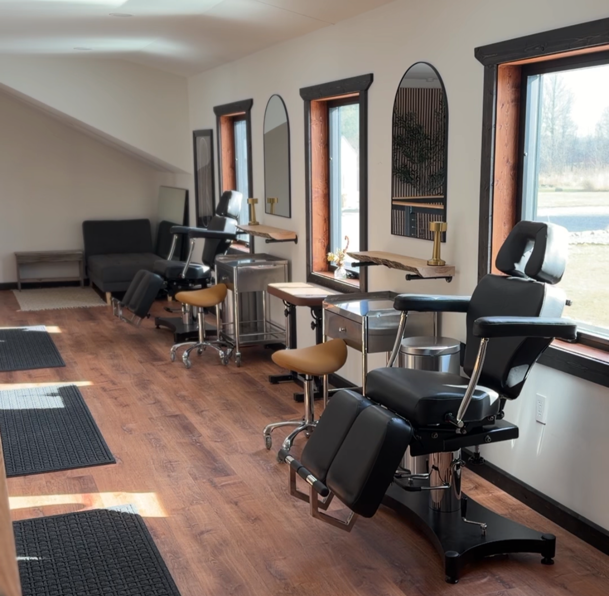 A clean, modern medical or dental treatment room with treatment chairs, stools, small tables, and decorative mirrors, illuminated by sunlight through large windows.