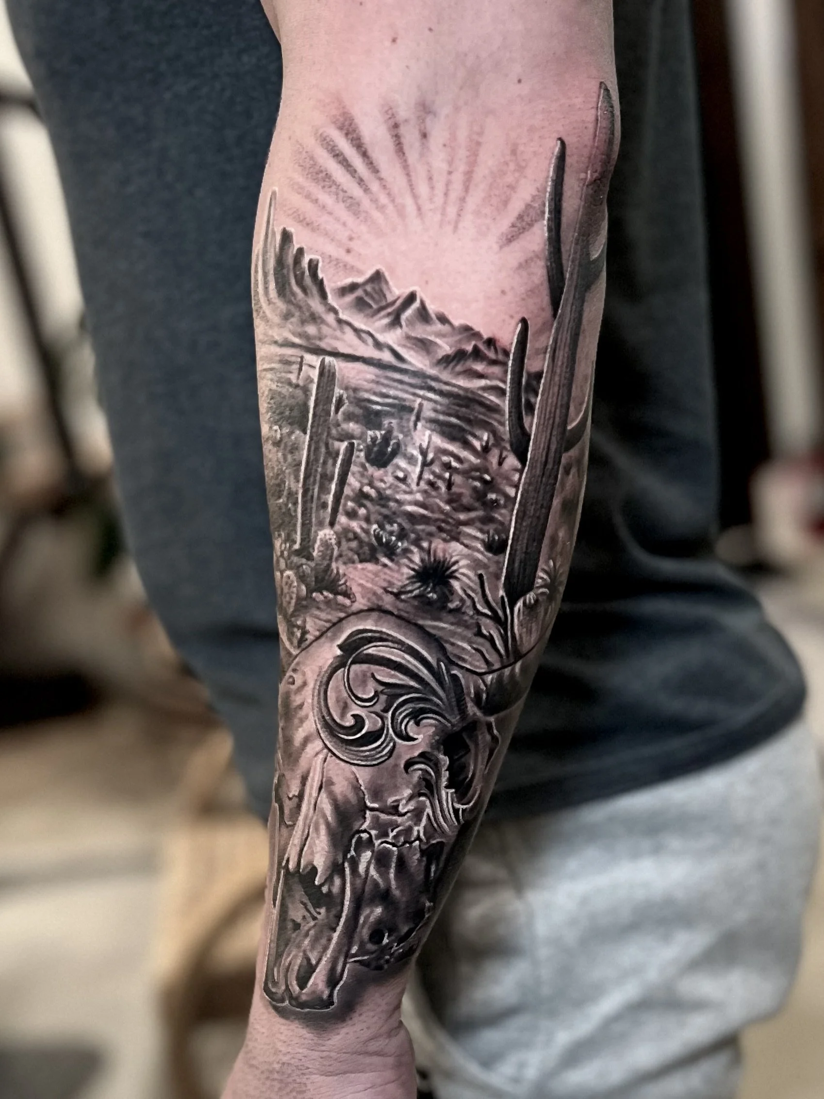 Detailed black and gray tattoo of a desert landscape with mountains, cacti, and a skull on a person's arm.