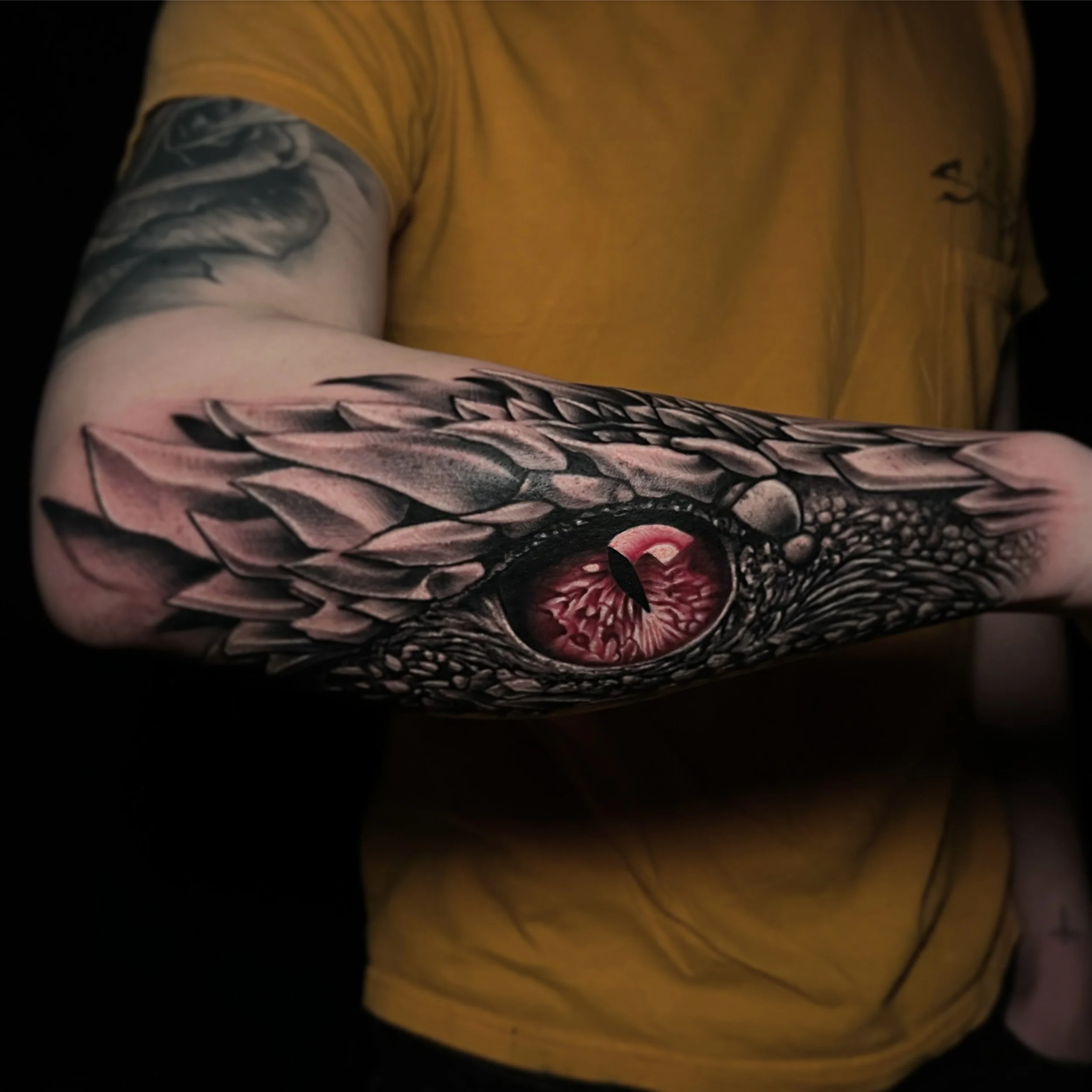 Tattoo of a dragon's eye on a person's arm, with detailed scales and feathers in black and gray shades.