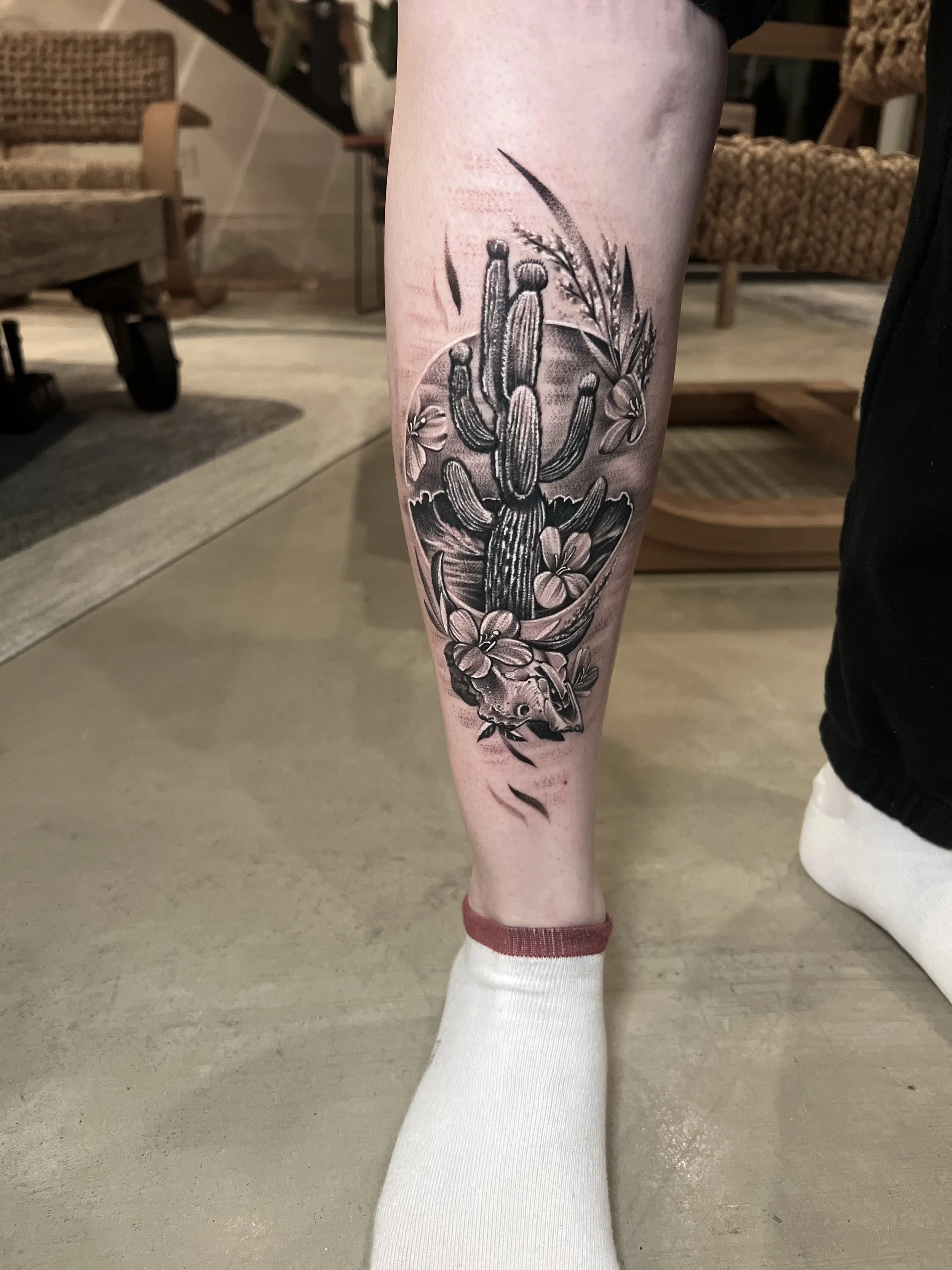 Black and gray tattoo on a person's calf featuring a desert scene with a cactus, a mouse skull, and floral elements.