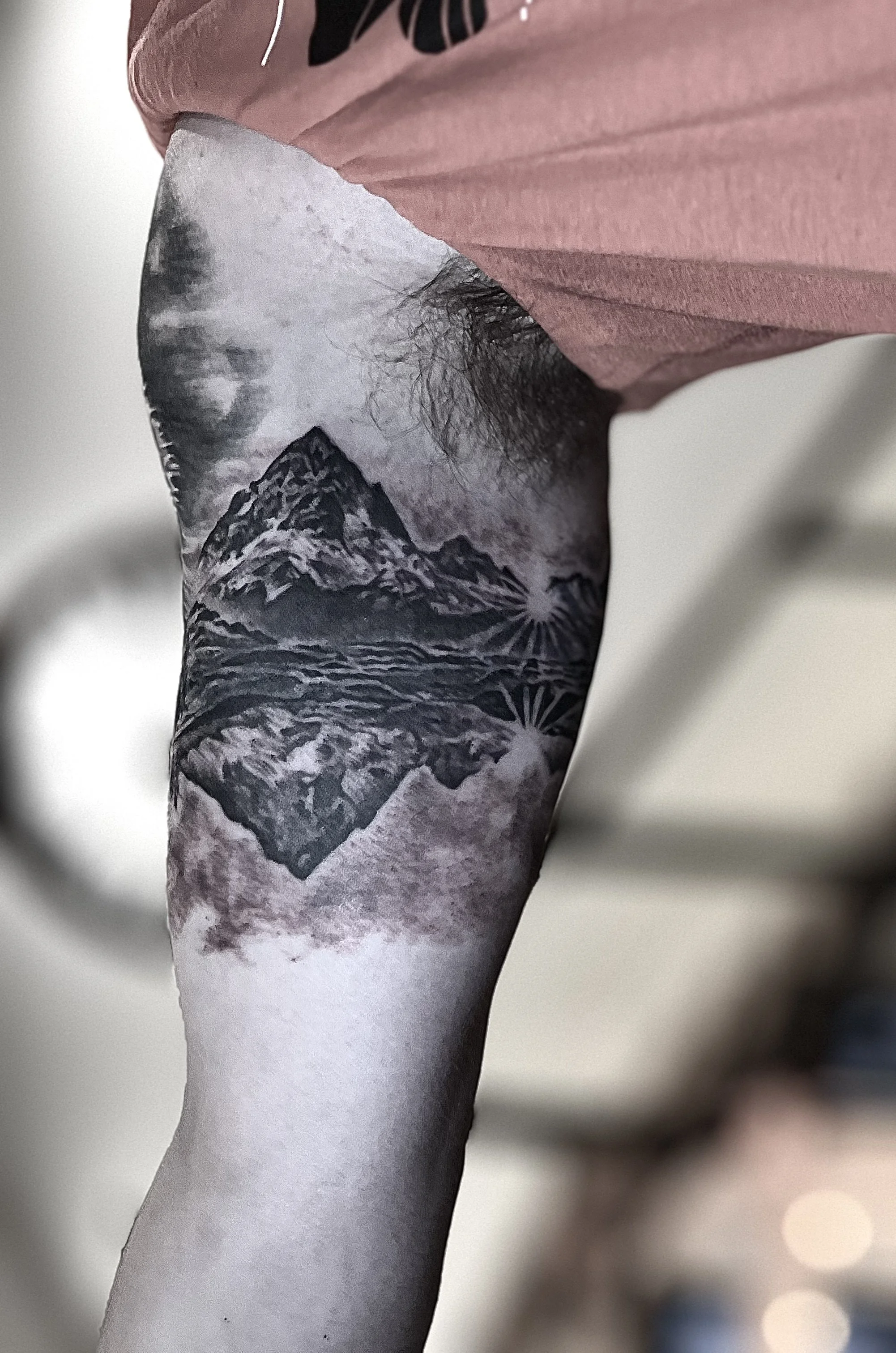 Tattoo of a mountain landscape with a lake, sun rays, and clouds on person's arm.