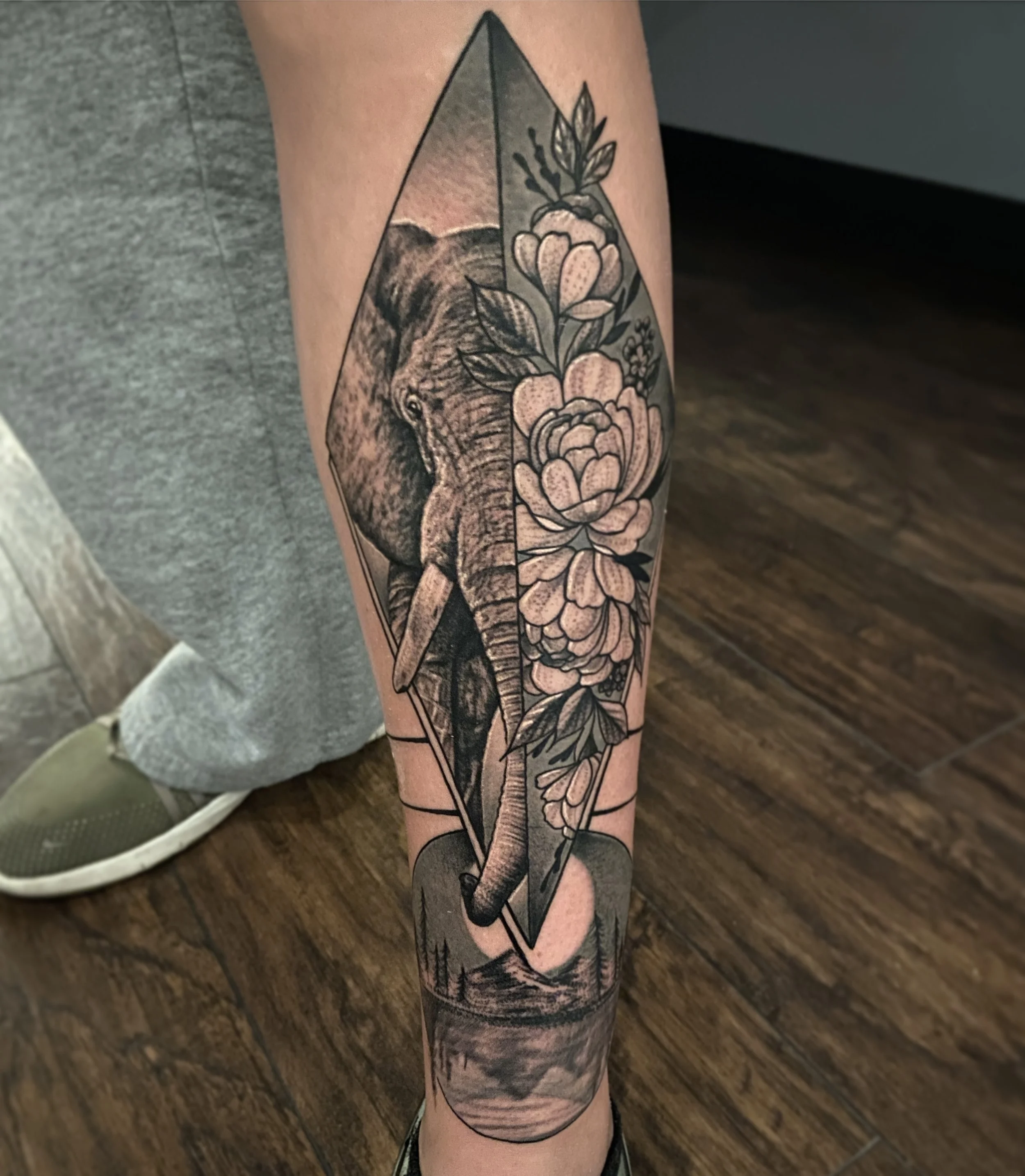 Tattoo of an elephant's head inside a geometric diamond shape, with detailed flowers and leaves behind it, on a person's forearm.