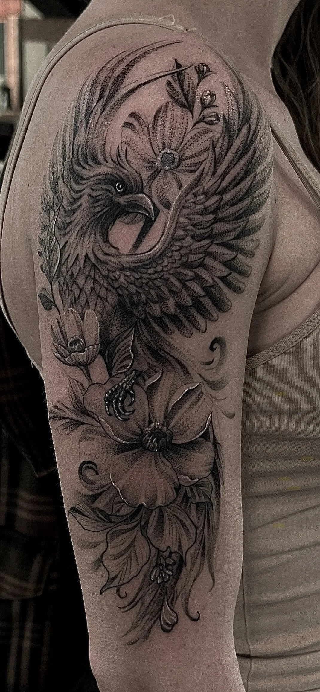 A detailed black and gray tattoo on a person's upper arm featuring an eagle with outstretched wings, surrounded by flowers and leaves.