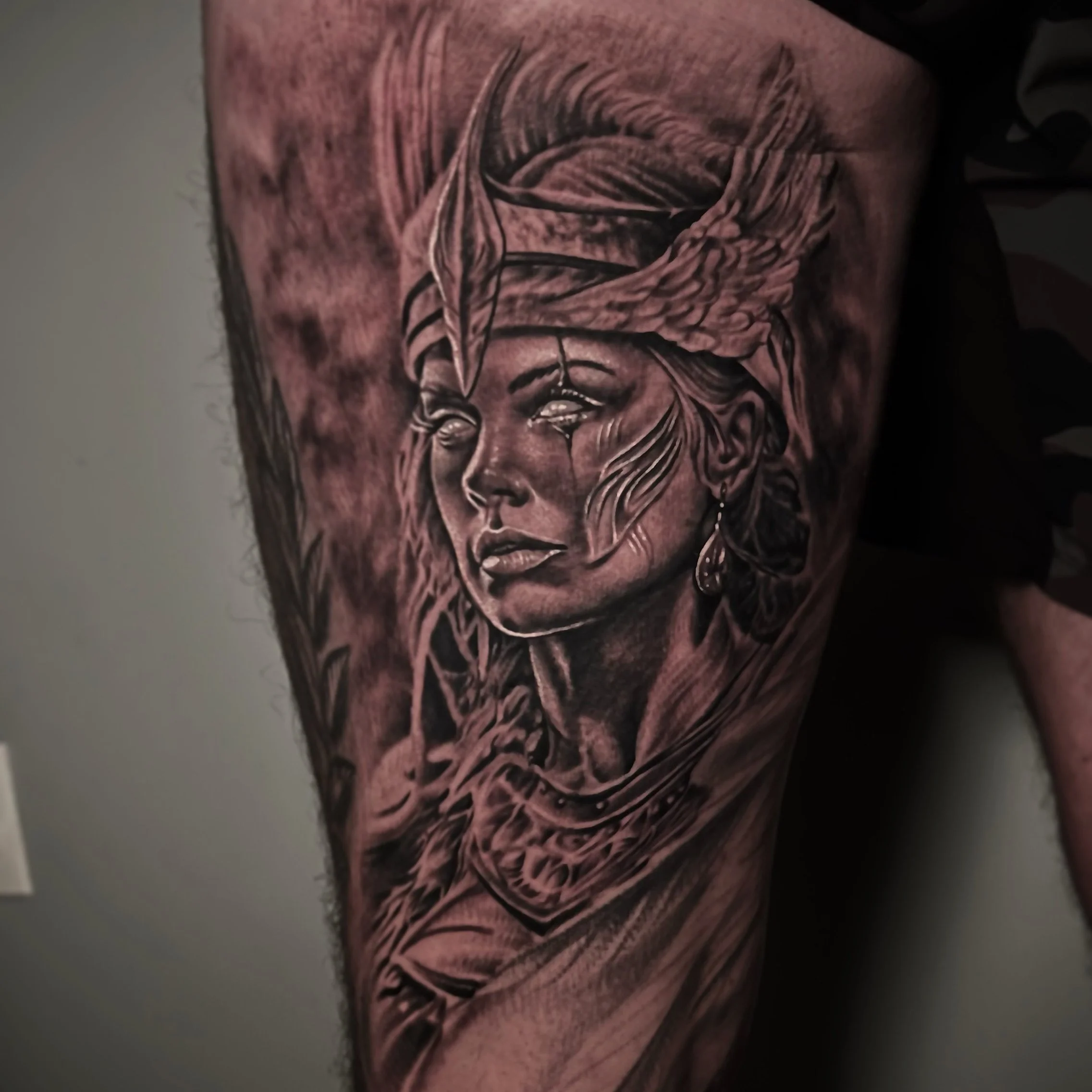 A detailed black and gray tattoo of a woman's face wearing a headscarf with feathers, earrings, and a necklace, on a person's upper arm.