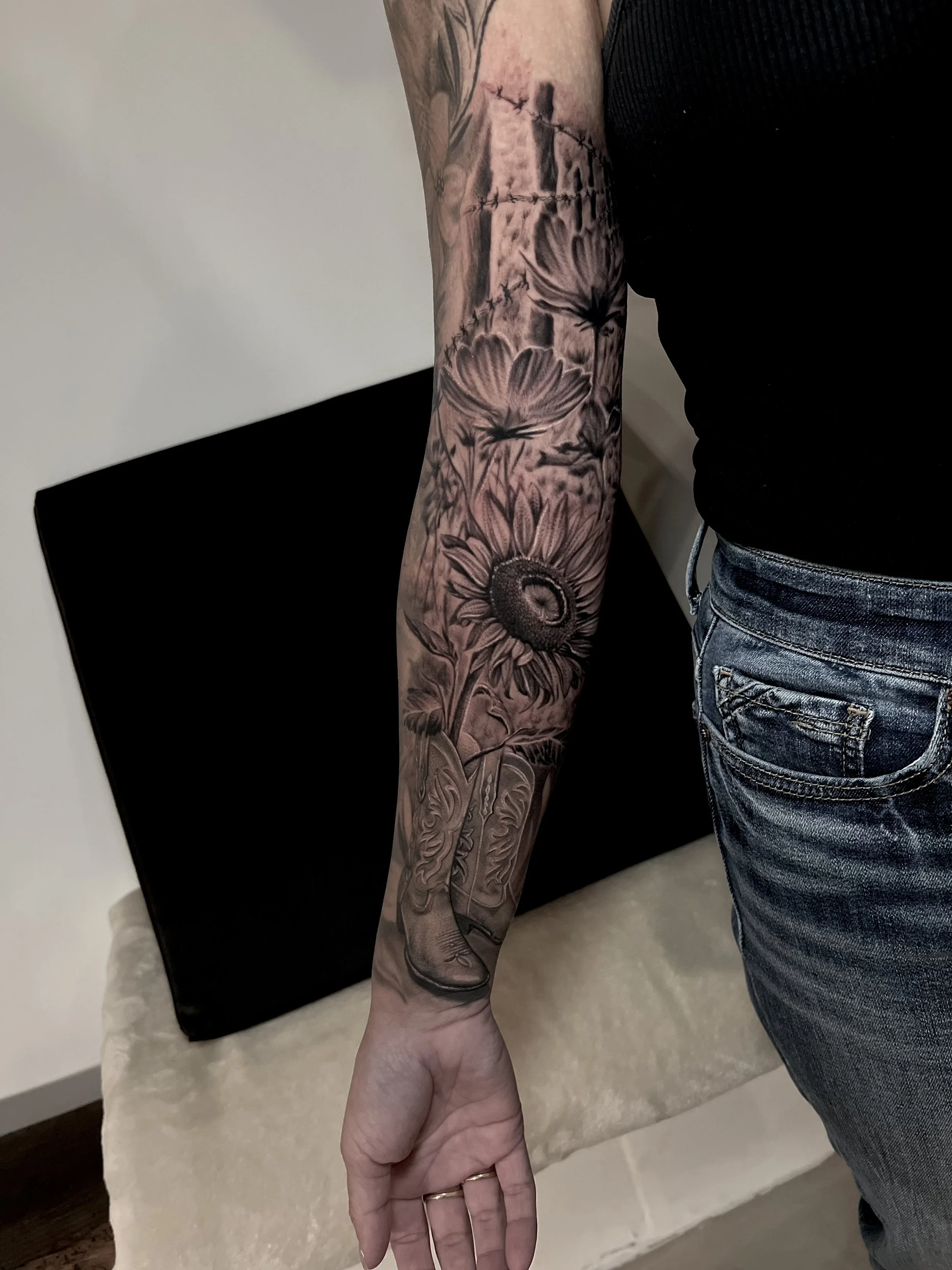 A person's arm with a detailed black and gray floral tattoo featuring daisies, a sunflower, and a cowboy boot, with the person wearing a black sleeveless top and blue jeans.