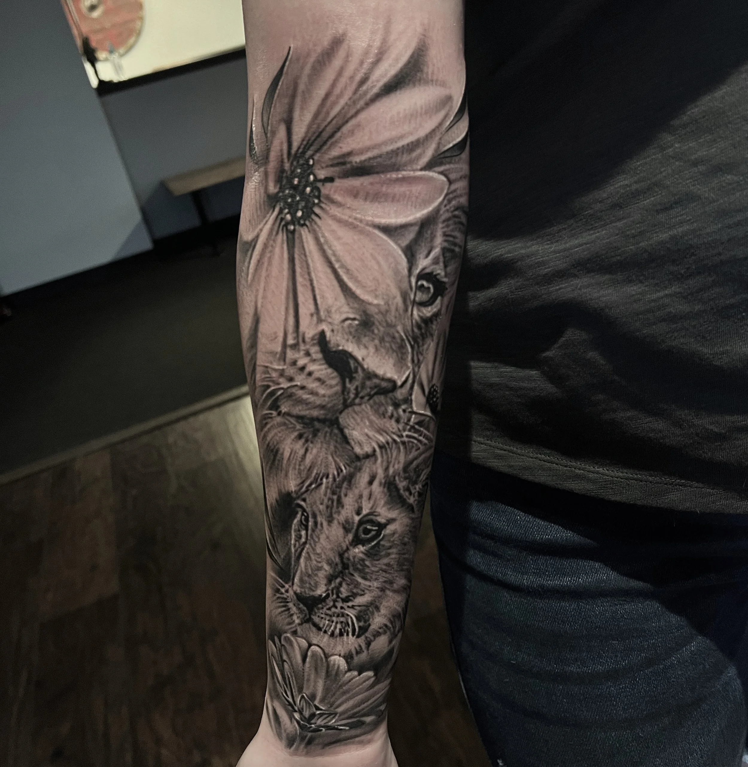 Black and grey tattoo of a lion's face surrounded by large flowers on a person's forearm.