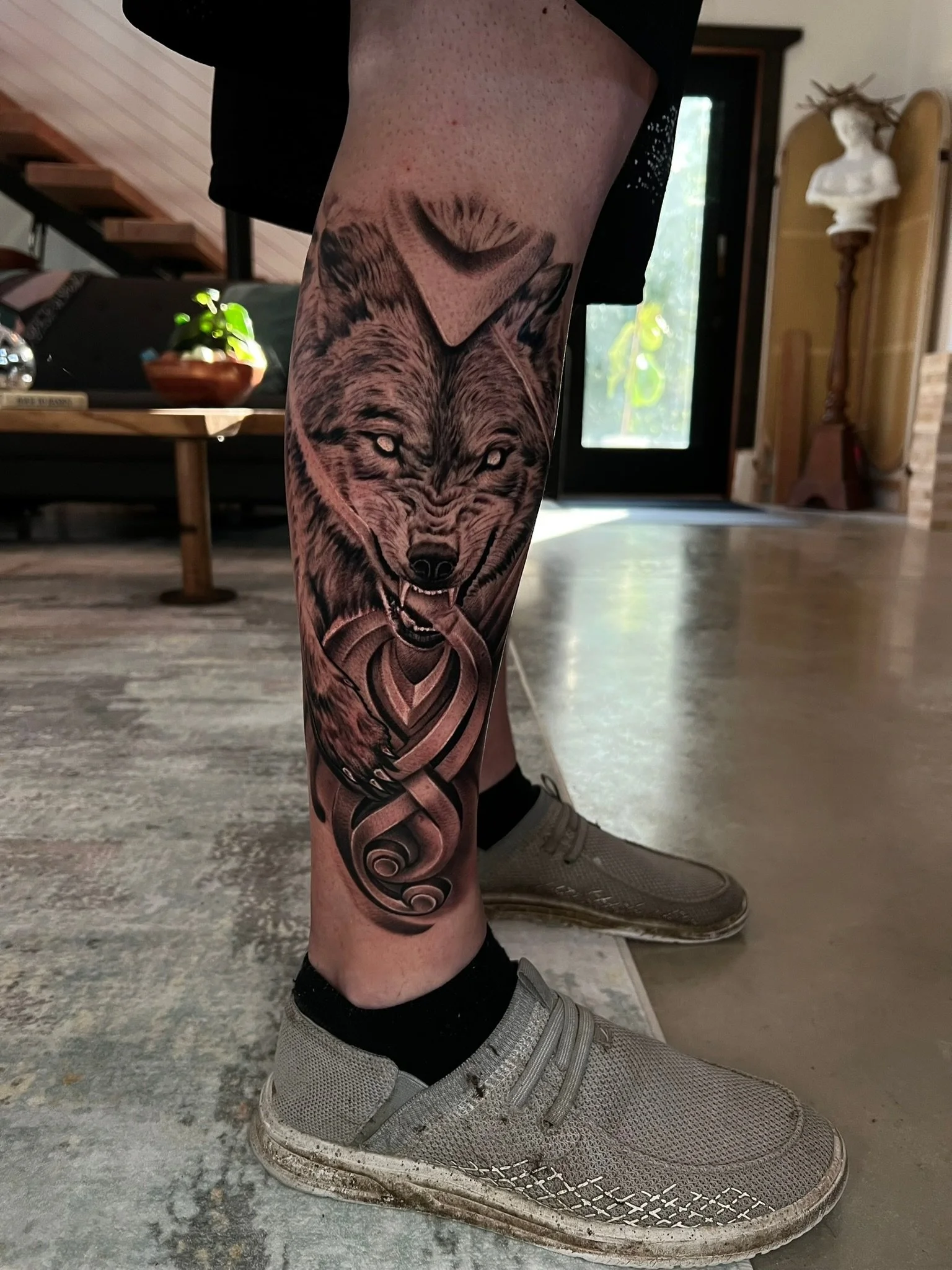 Tattoo of a wolf with an intricate design on a person's calf, with a wooden floor and home interior in the background.