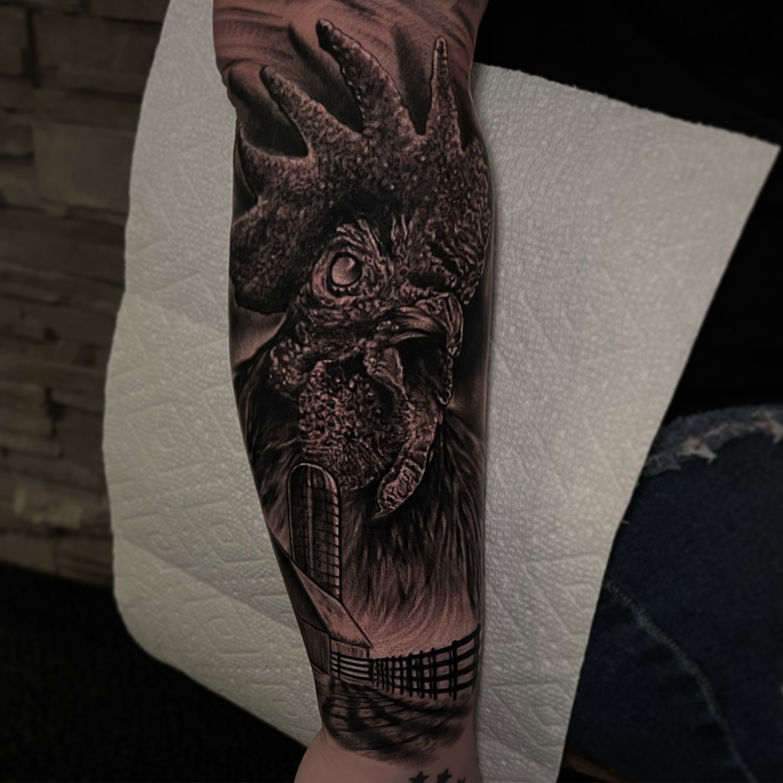Black and gray tattoo of an owl with a deer head on a person's forearm, with a tree, a window, and a fence in the background.