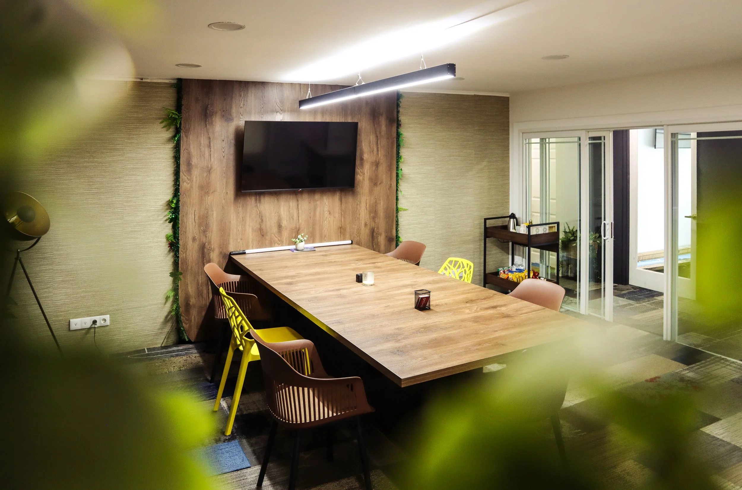 Modern dining room with a long wooden table and various colored chairs, a wall-mounted flat-screen TV, a sliding glass door, and decorative green plants along the wall.