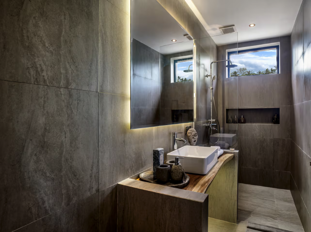 Modern bathroom with a large illuminated mirror, a white vessel sink on a wooden vanity, decorative items, and a walk-in shower with a rainfall showerhead, small windows, and recessed shelf.