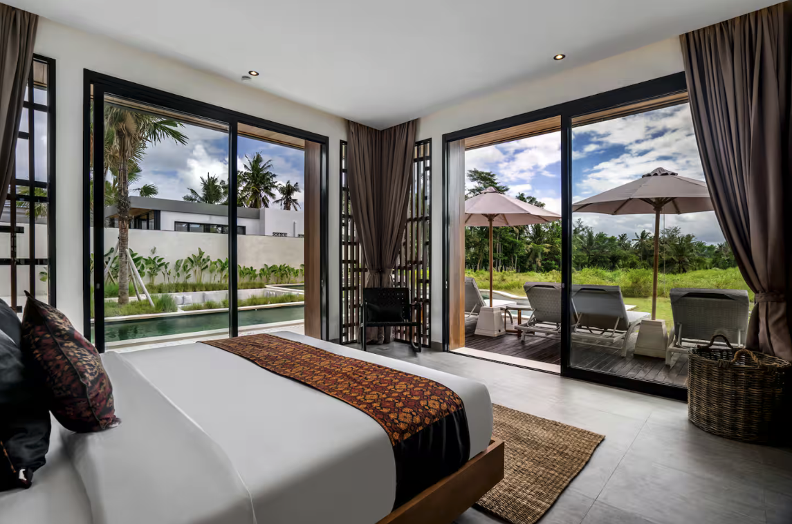 Modern bedroom with large sliding glass doors opening to a patio with lounge chairs and umbrellas, tropical trees, and a pool outside.