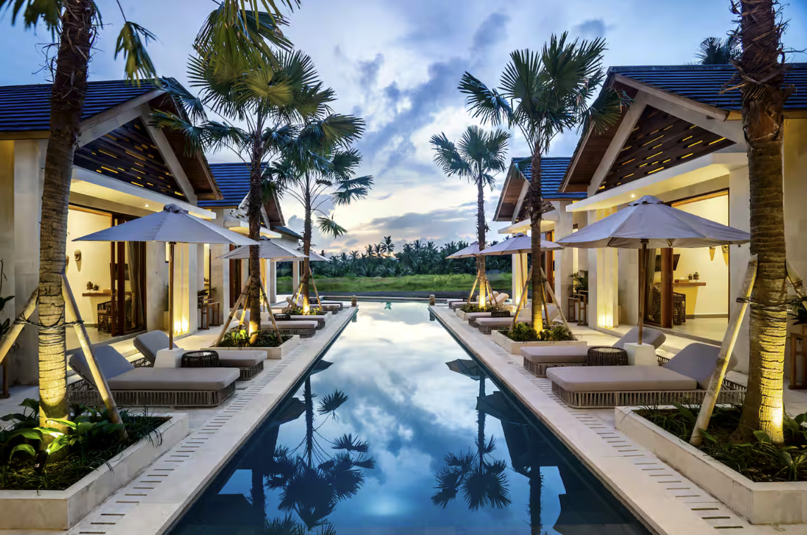 View of a luxurious infinity pool with lounge chairs and umbrellas, surrounded by palm trees and tropical landscaping, overlooking a lush green landscape at sunset.