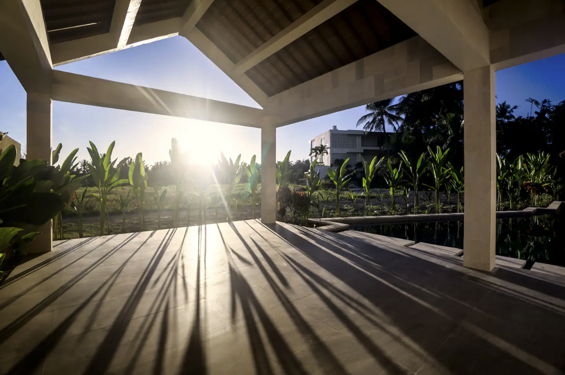 Sunshine shining through an open porch with shadows cast on the floor, overlooking a garden with green plants and palm trees, and a modern house in the background.