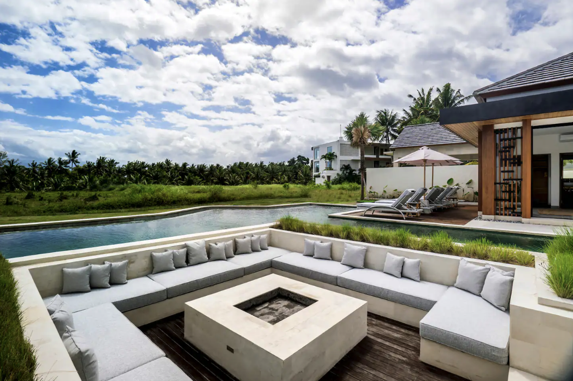 Luxury backyard with a pool, outdoor seating area with cushions, lounge chairs under an umbrella, and a view of palm trees under a partly cloudy sky.